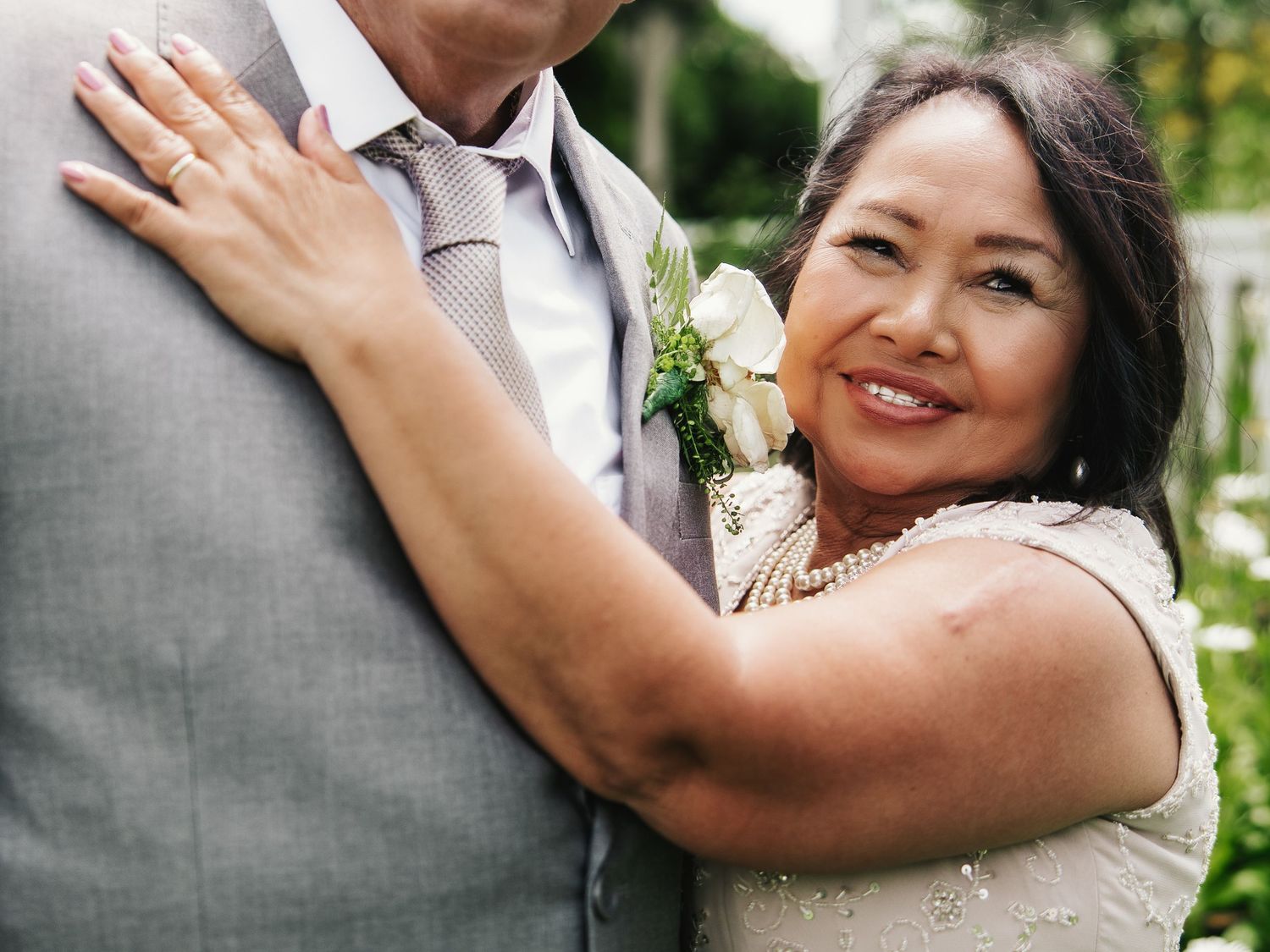 Smiling woman in beaded dress embraces man in gray suit with white boutonniere at an outdoor wedding ceremony.