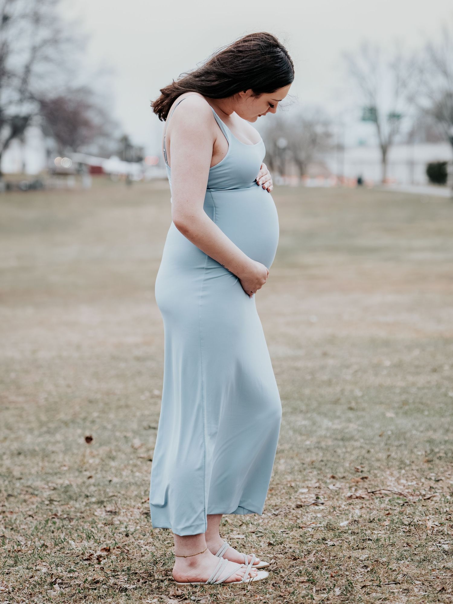 Pregnant woman in a light blue maternity dress cradling her baby bump while looking down in an outdoor park setting.