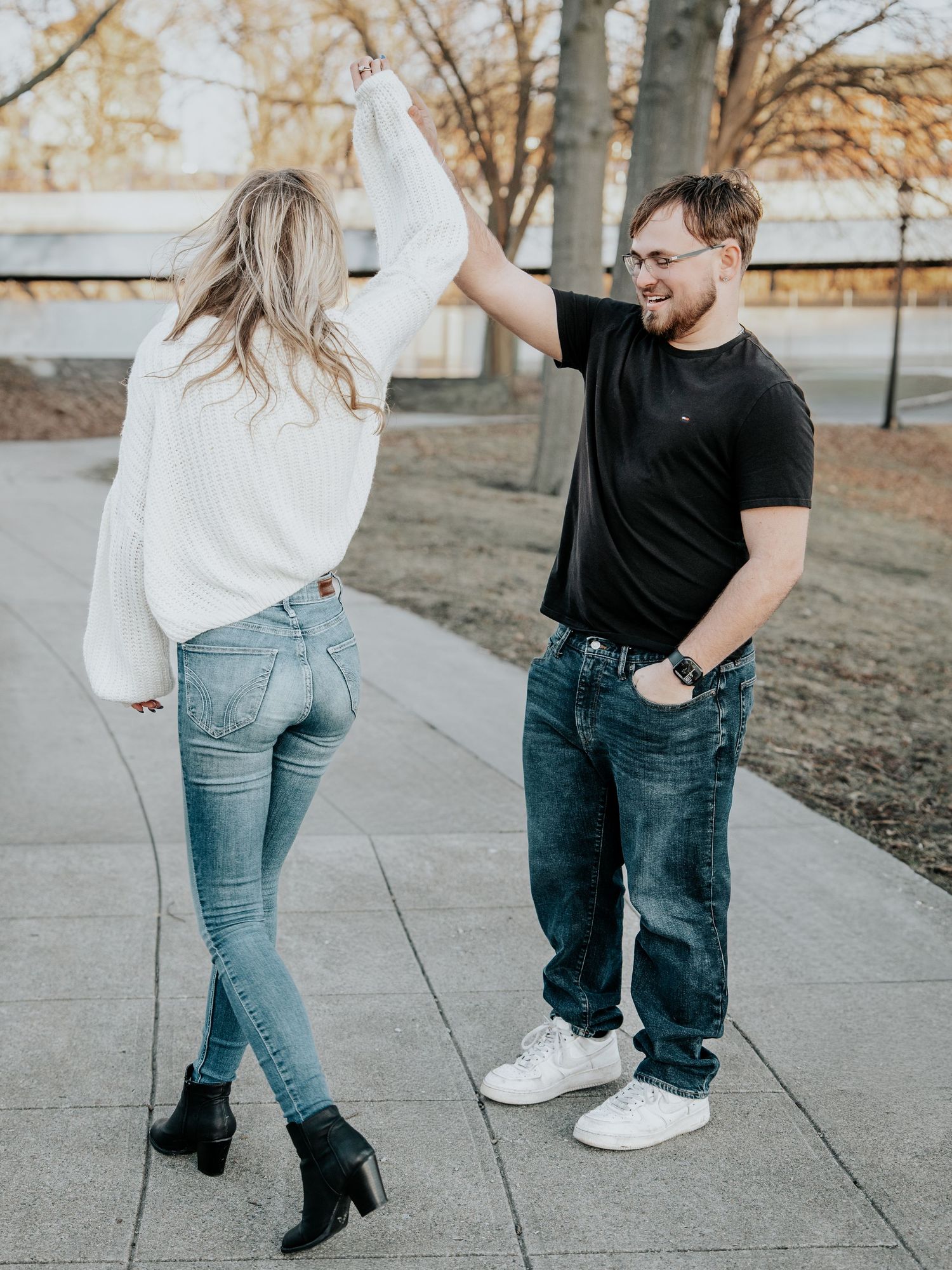 Couple dancing outdoors on a paved path, woman in white sweater and jeans, man in black tshirt and jeans, trees in background.