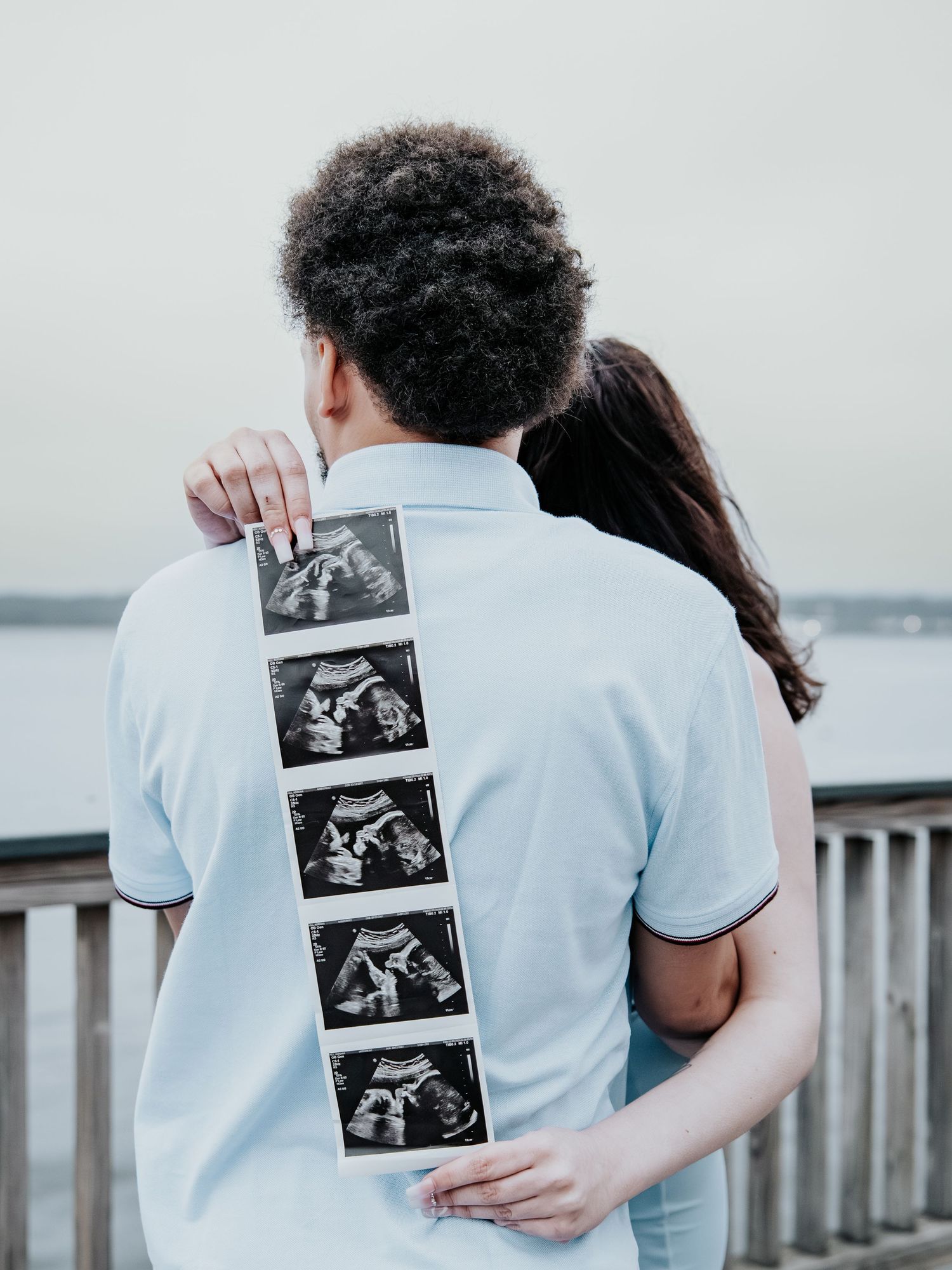 Couple embracing on a dock holding ultrasound photos behind their backs for a pregnancy announcement.