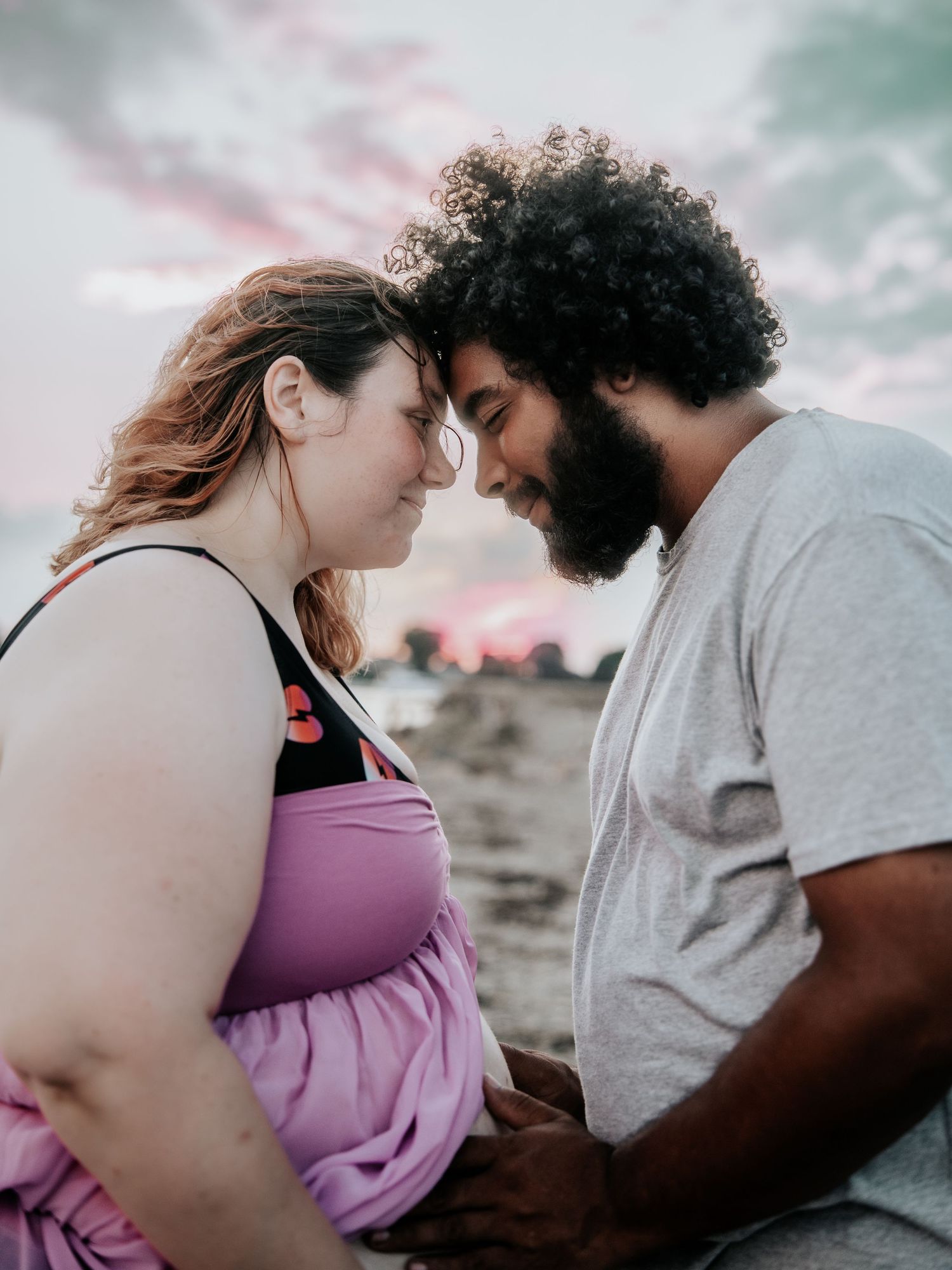 A couple shares a romantic forehead touch on the beach at sunset, the woman in purple and man in gray.