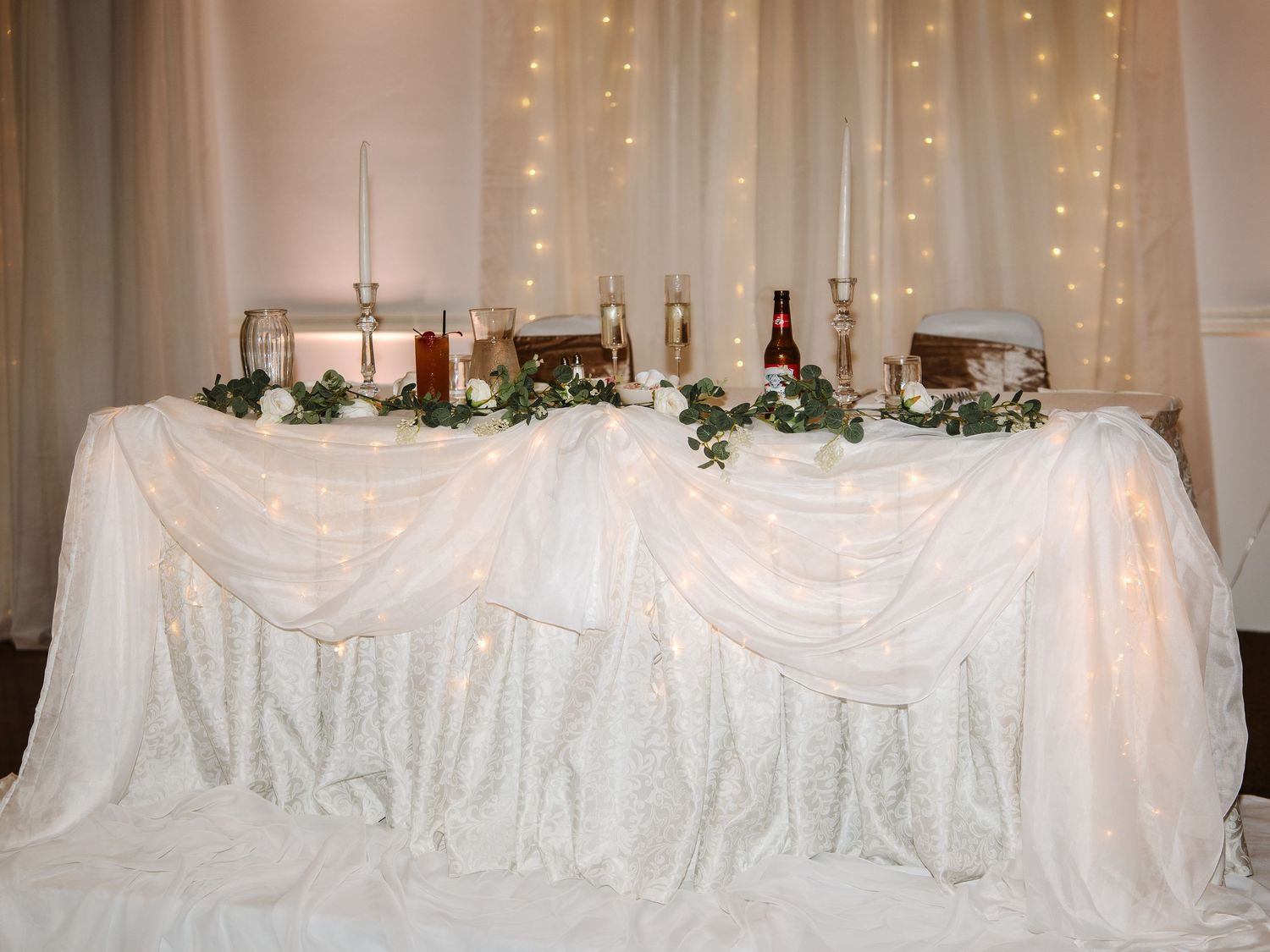 Elegant wedding head table with white draped fabric, fairy lights backdrop, greenery garland, and candles.