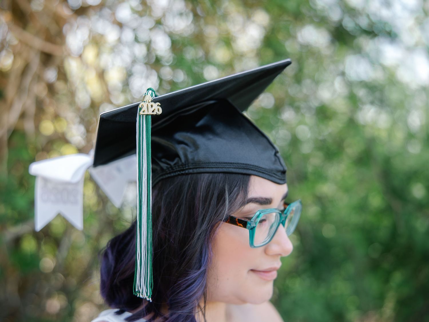 Close-up of graduate wearing black mortarboard cap with green tassel and teal glasses outdoors.