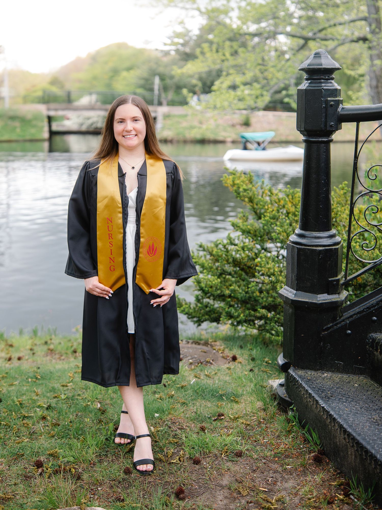 Nursing graduate in black cap and gown with gold honor stole posing by a scenic waterfront.