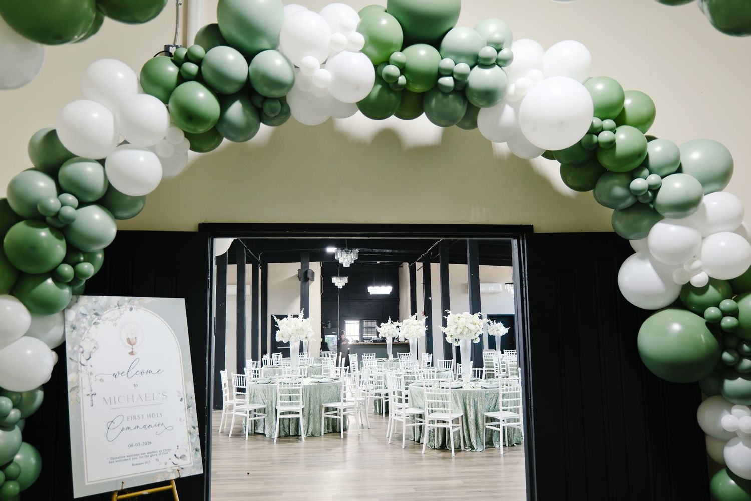 Sage green and white balloon arch framing entrance to elegant baptism reception hall with floral centerpieces.