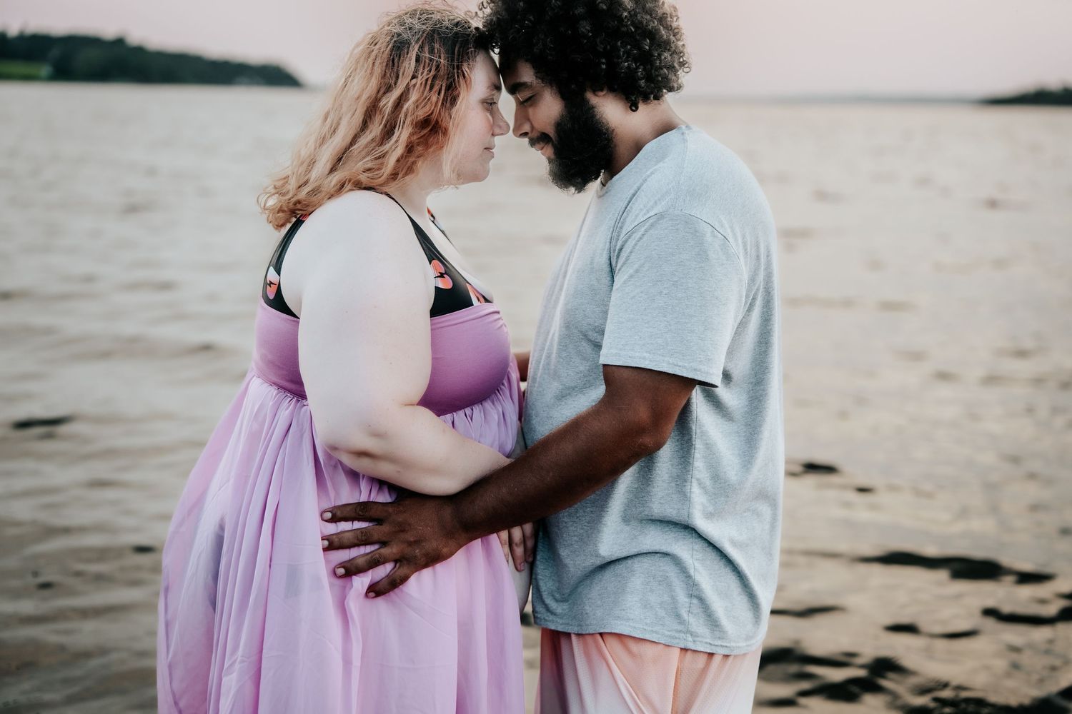 Pregnant couple sharing an intimate moment on a beach at sunset, woman in purple dress, man in gray shirt.