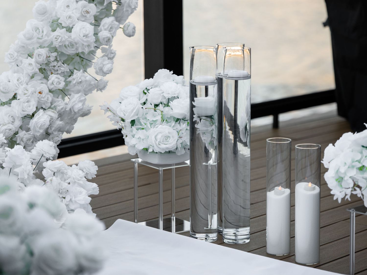 Elegant white wedding table decor with glass cylinder vases, white roses, and lit candles for a romantic event.