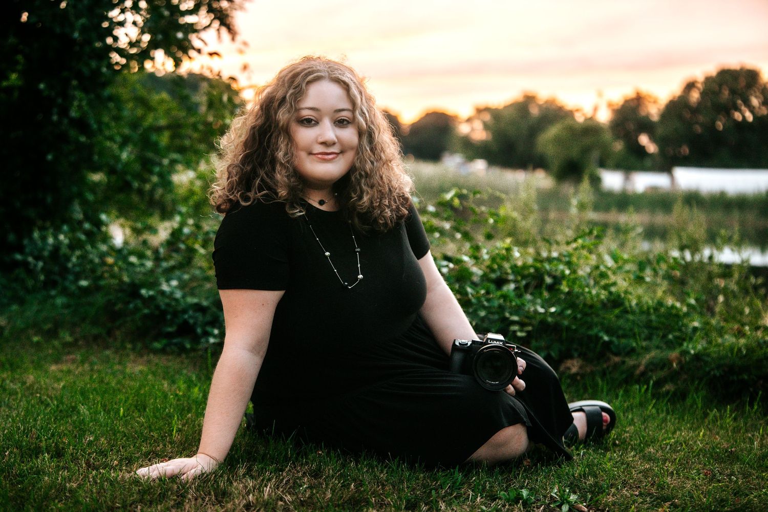Curly-haired woman sitting on grass holding a camera at sunset near lush greenery and a lake.
