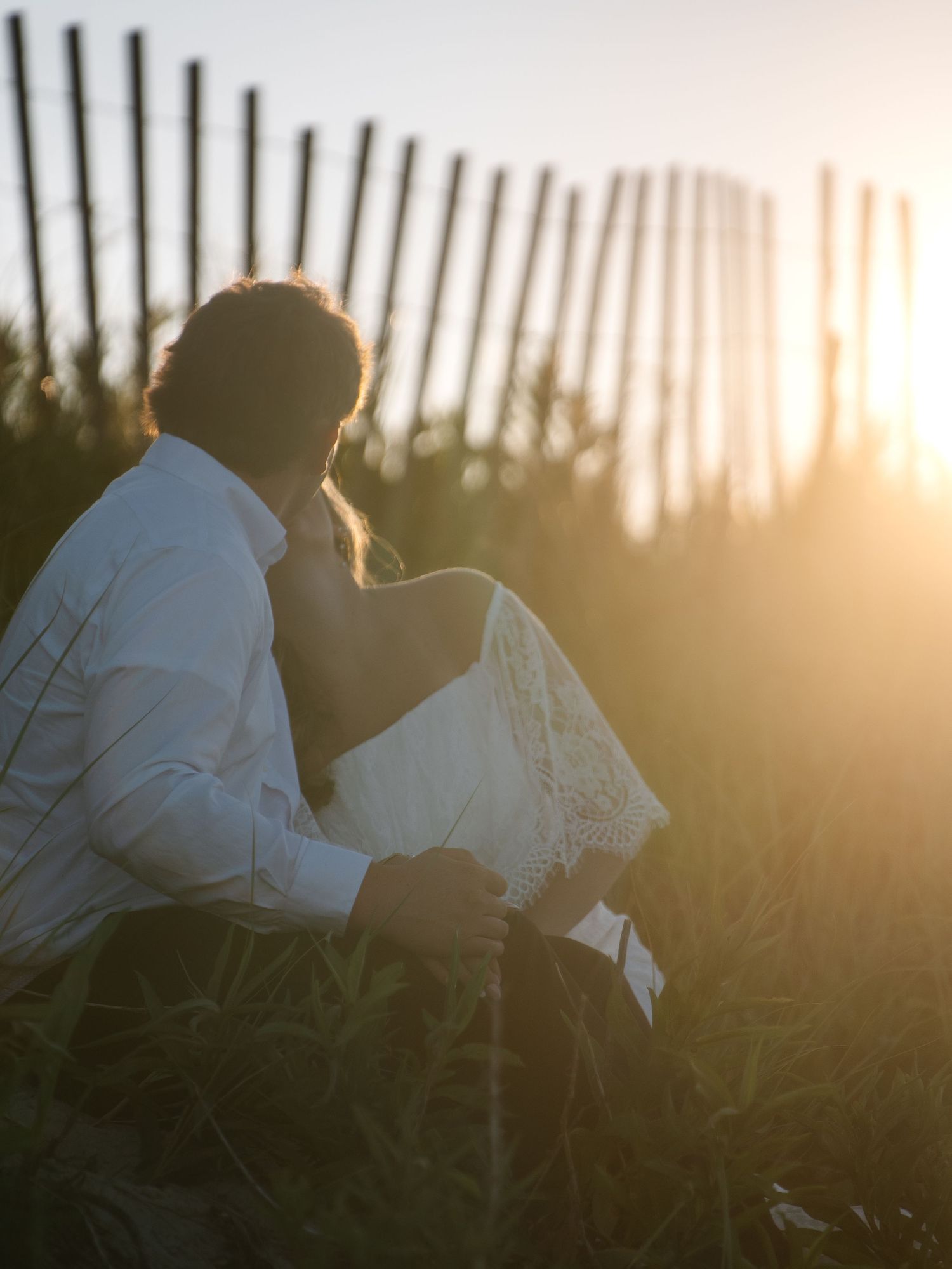 Couple sitting intimately together in tall grass at sunset, viewed from behind near a wooden fence.