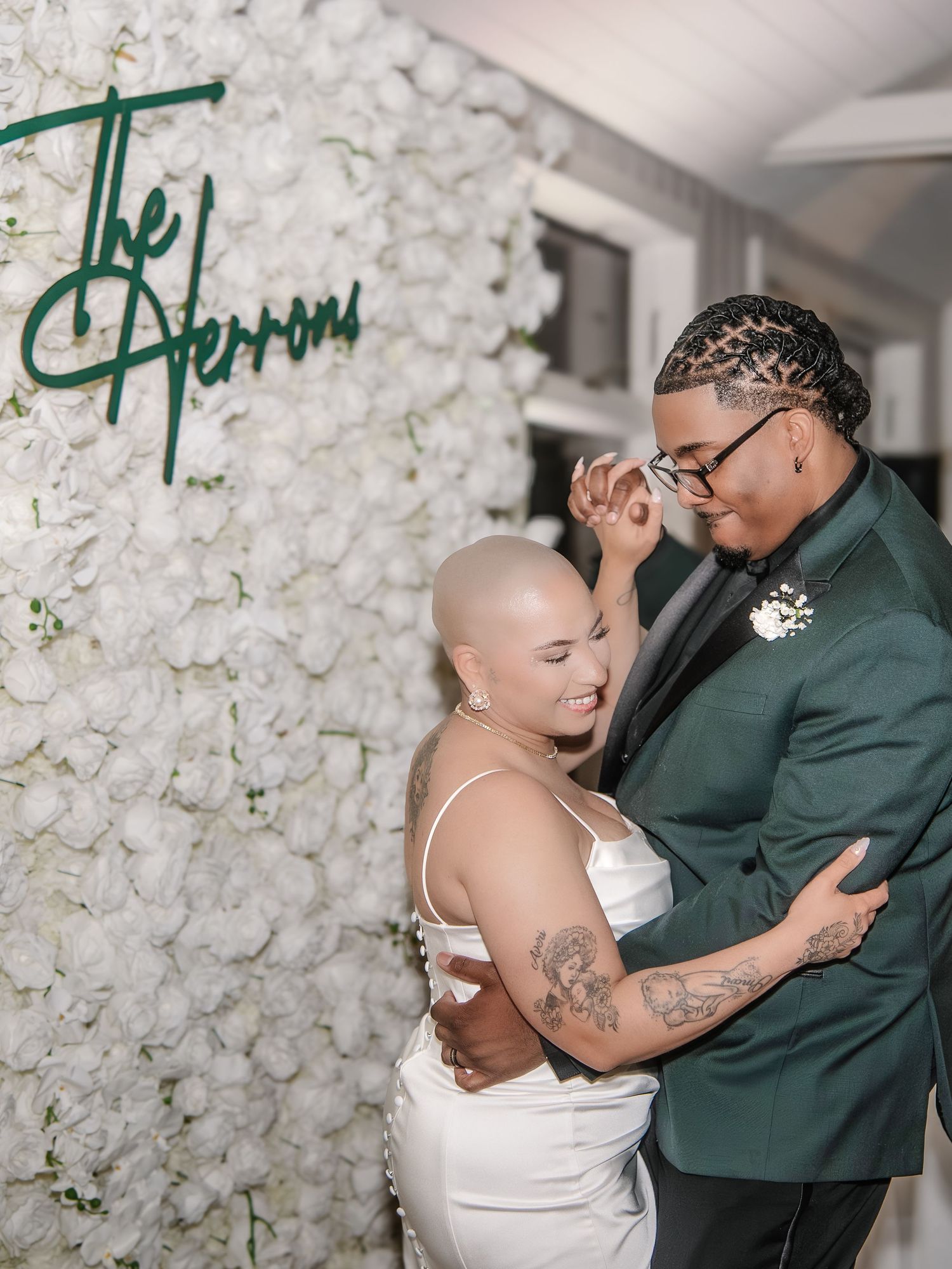 Two brides share a romantic dance in front of a white floral wall with green neon sign reading The Herrons.
