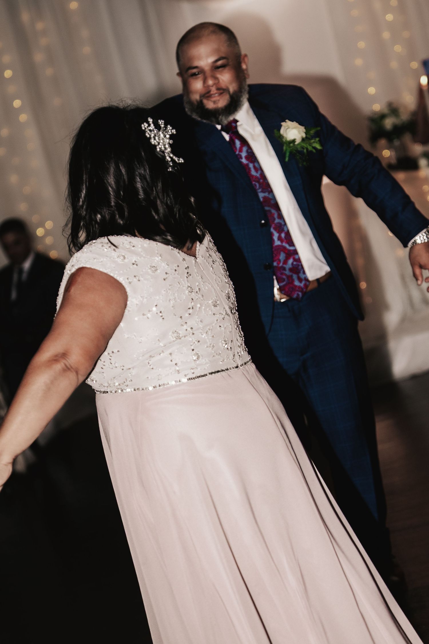 A couple shares a joyful first dance, bride in beaded gown and groom in navy suit with floral tie at wedding reception.