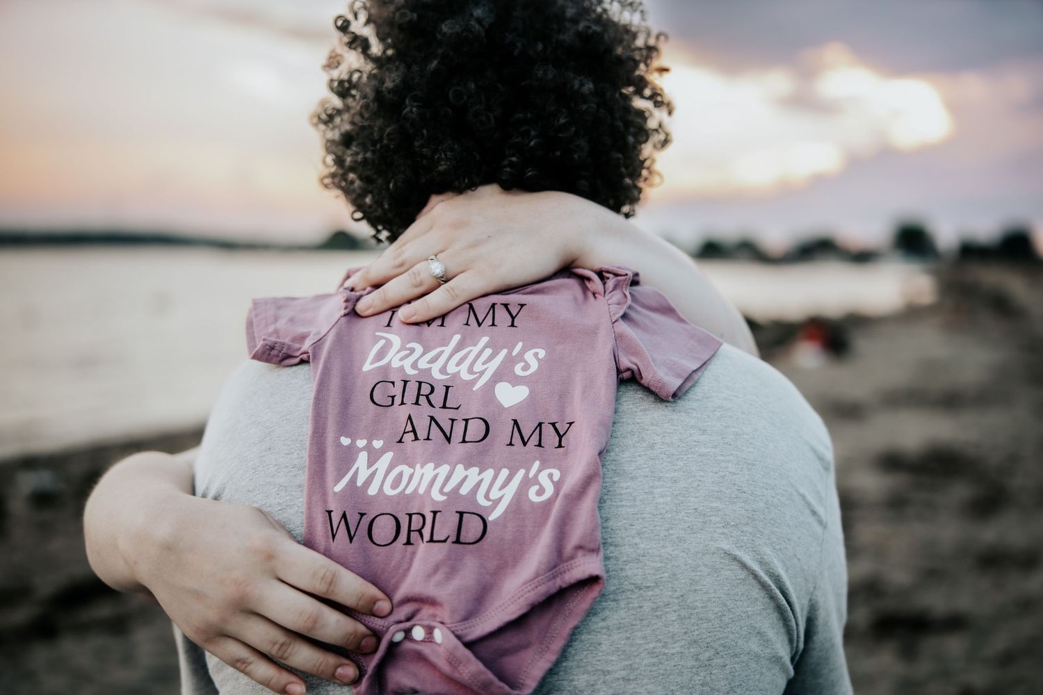 Parents holding baby in pink onesie reading Daddy's Girl and Mommy's World at sunset beach.