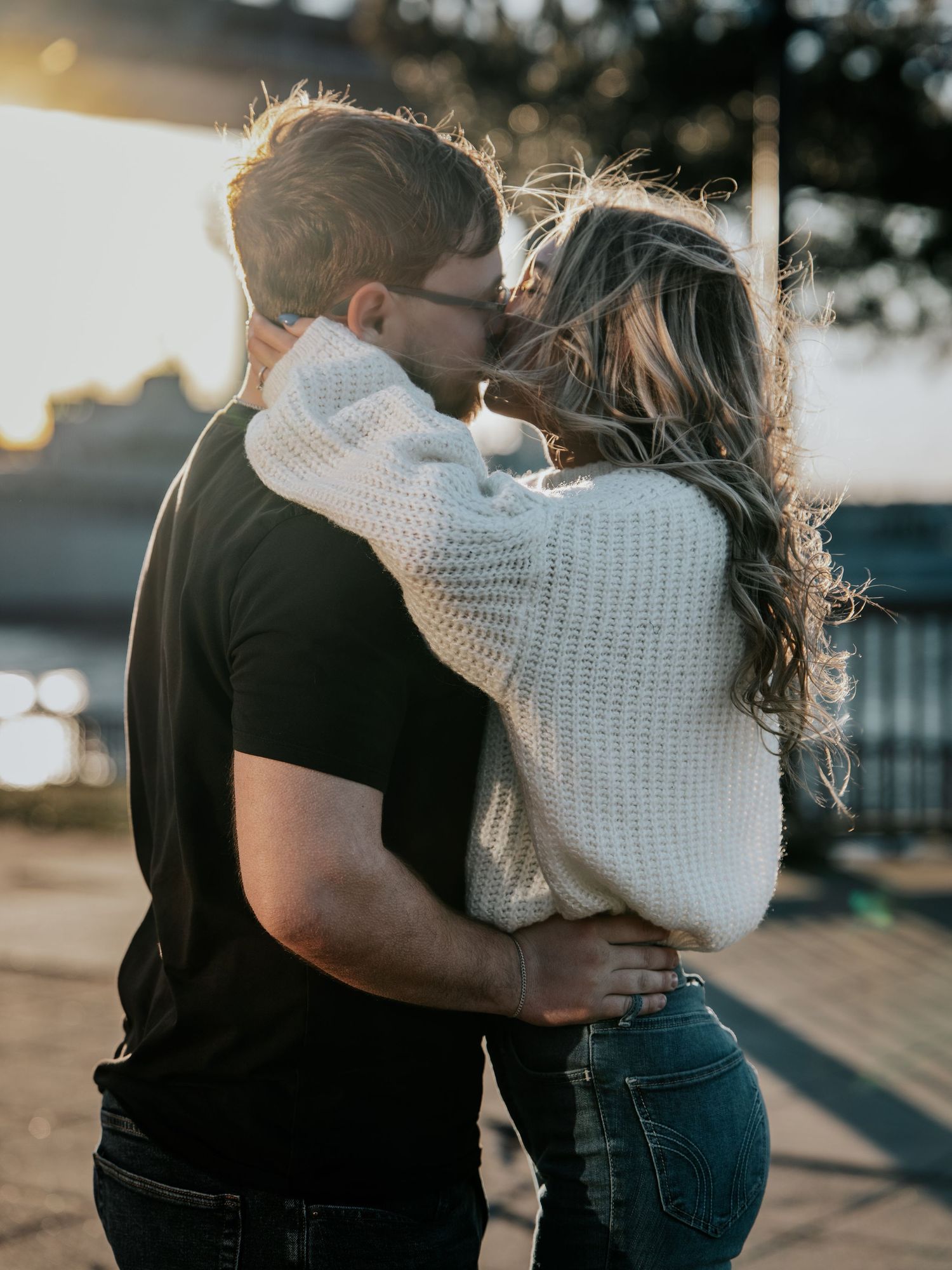 Couple sharing a romantic kiss at sunset, woman in white knit sweater and jeans embracing man in black t-shirt outdoors.
