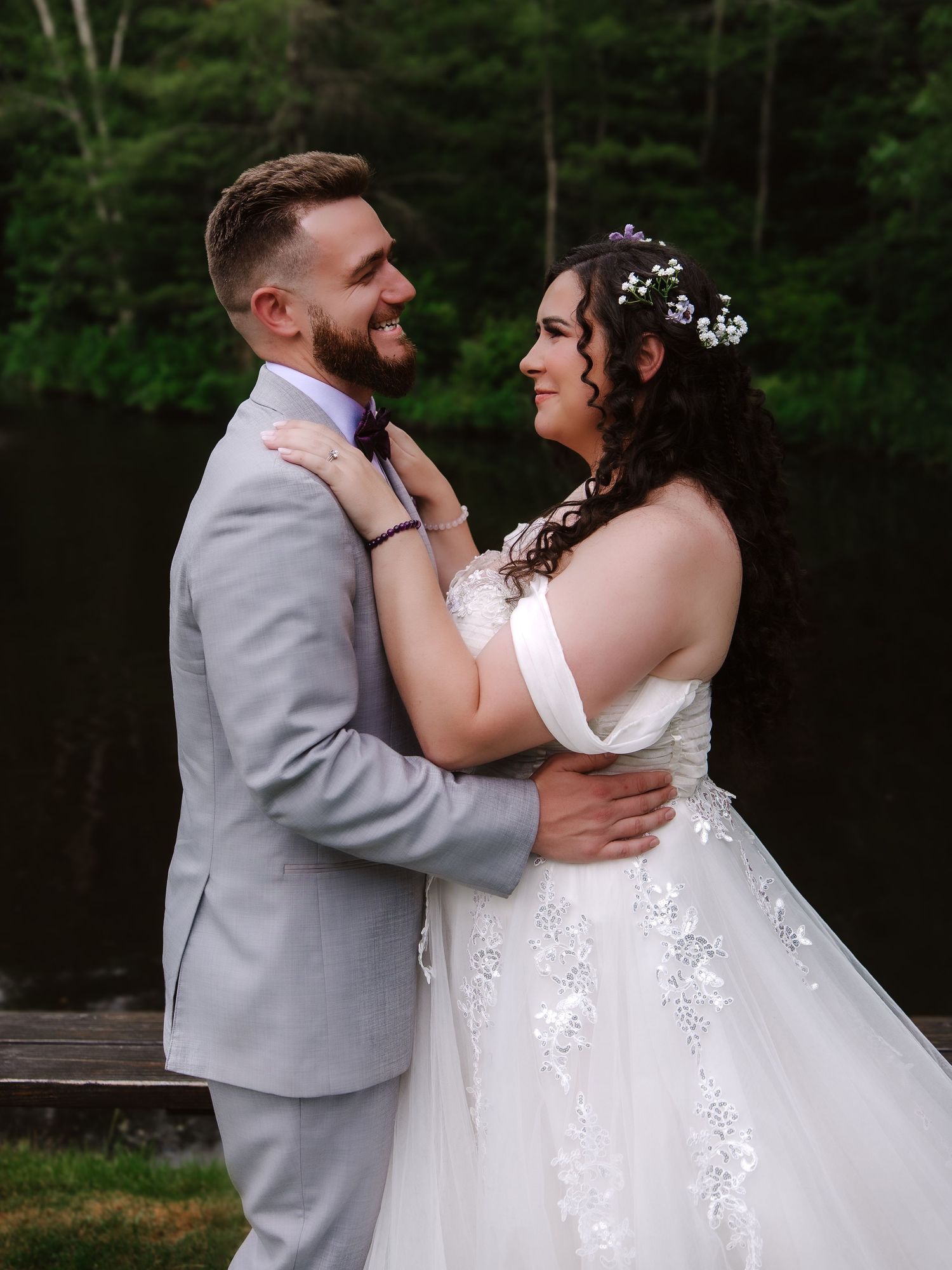 Smiling bride and groom embracing near a lake, she in a lace wedding gown and he in a grey suit.