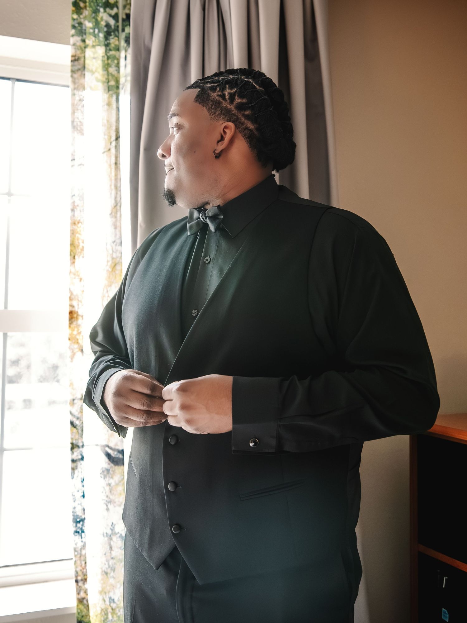 Man in dark green dress shirt and bow tie buttoning up near a window, preparing for a formal event.