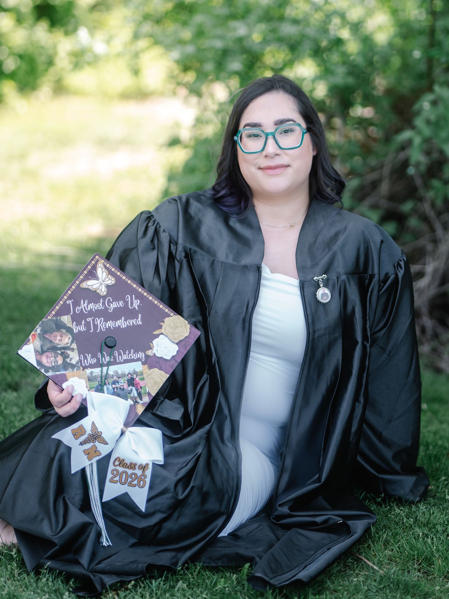 Graduate in black gown sitting on grass holding decorated mortarboard cap reading I Almost Gave Up.