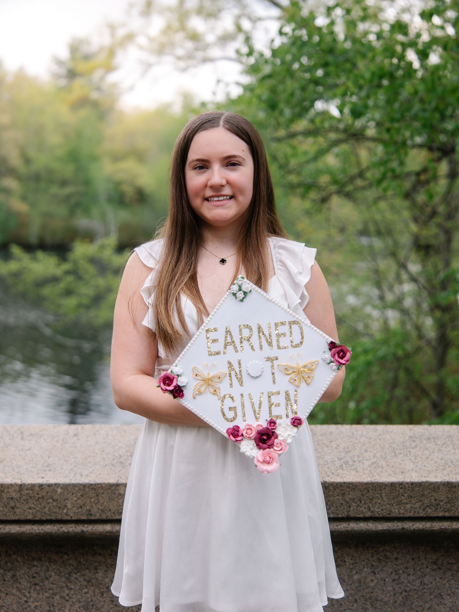 Smiling graduate in white dress holding decorated mortarboard cap reading Earned Not Given near a lake.
