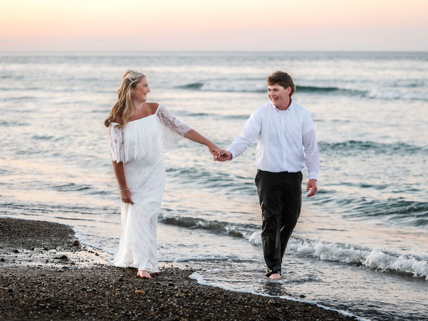 Bride in white off-shoulder gown and groom holding hands walking along ocean shoreline.