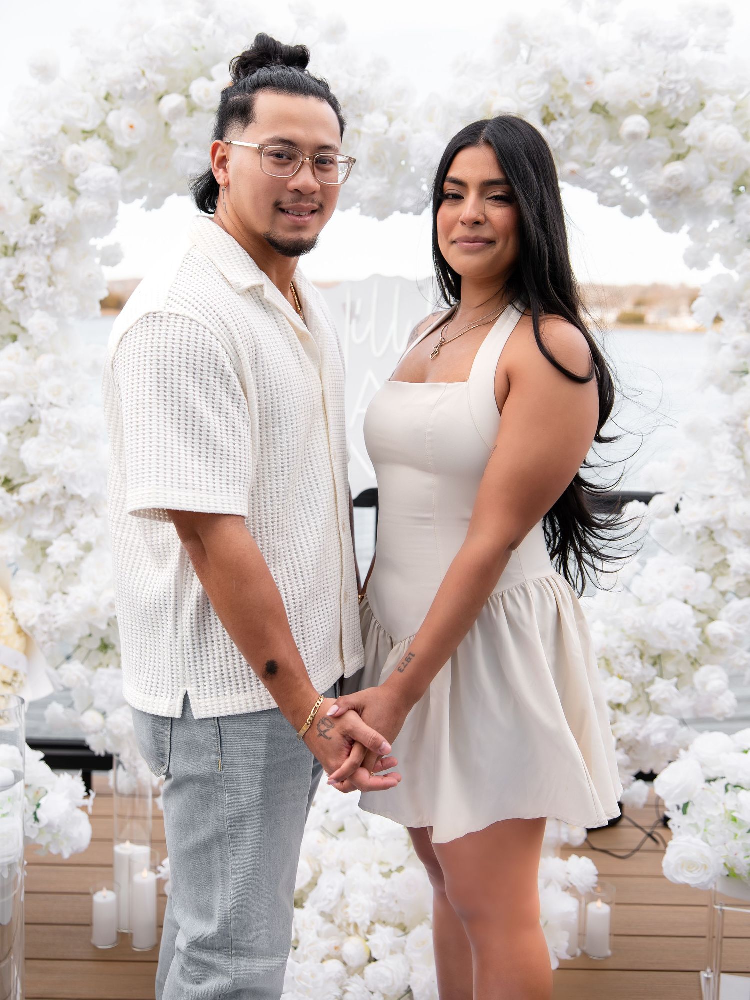 Engaged couple posing hand in hand before a stunning white floral arch near the waterfront.