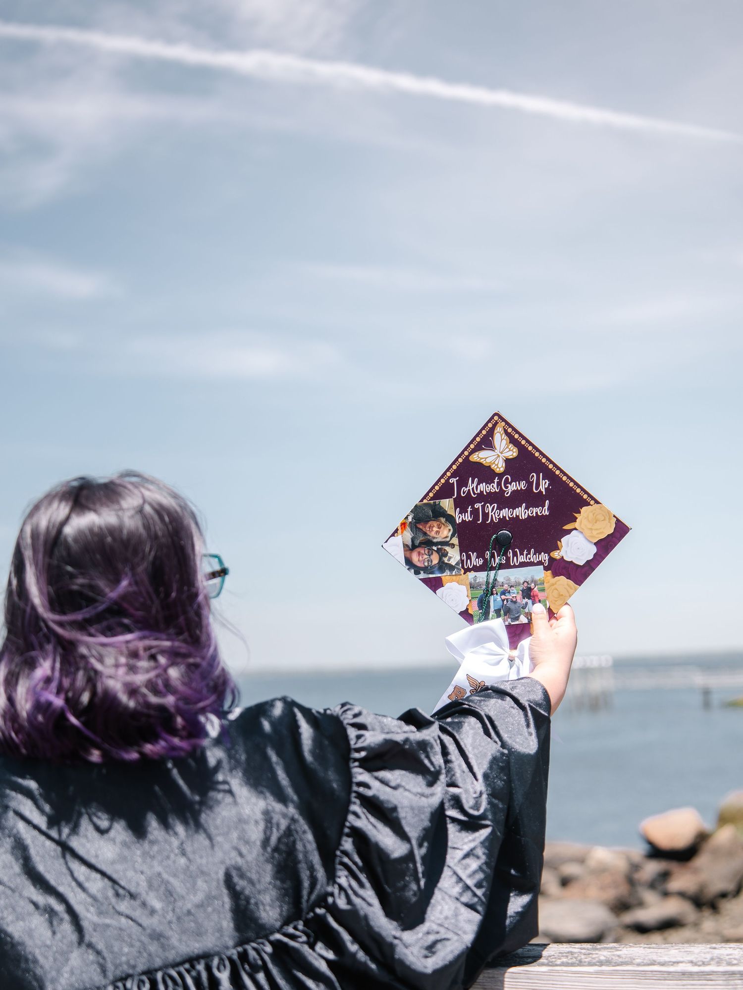 Graduate holding decorated purple mortarboard cap overlooking ocean on sunny graduation day.