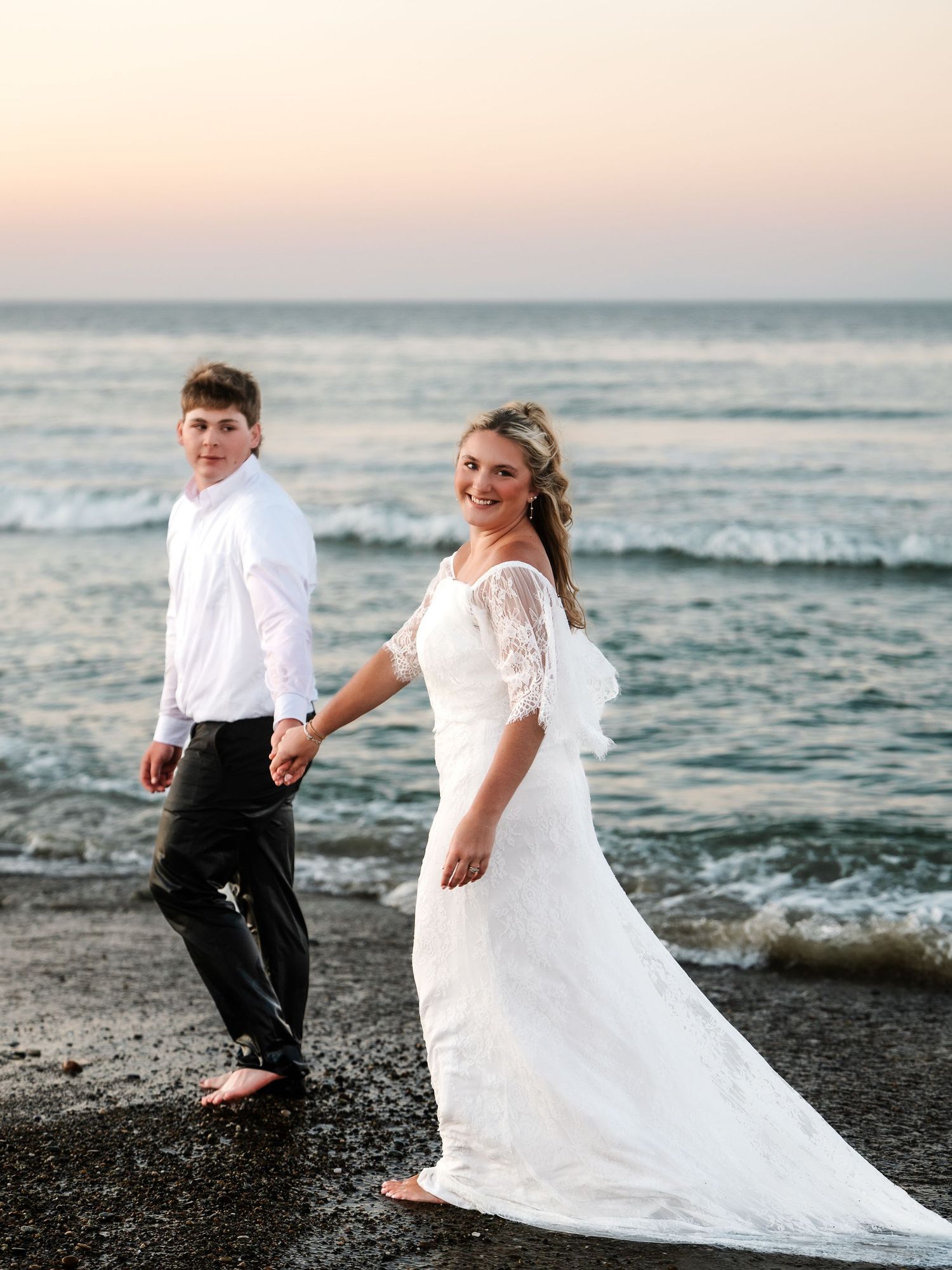 Bride in white lace gown and groom in white shirt walking barefoot on beach at sunset.