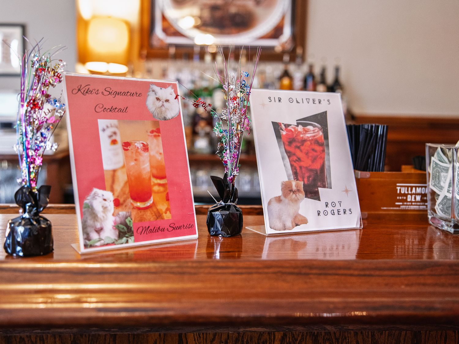 Two personalized wedding signature cocktail menu cards displayed on a wooden bar with decorative sparkle arrangements.