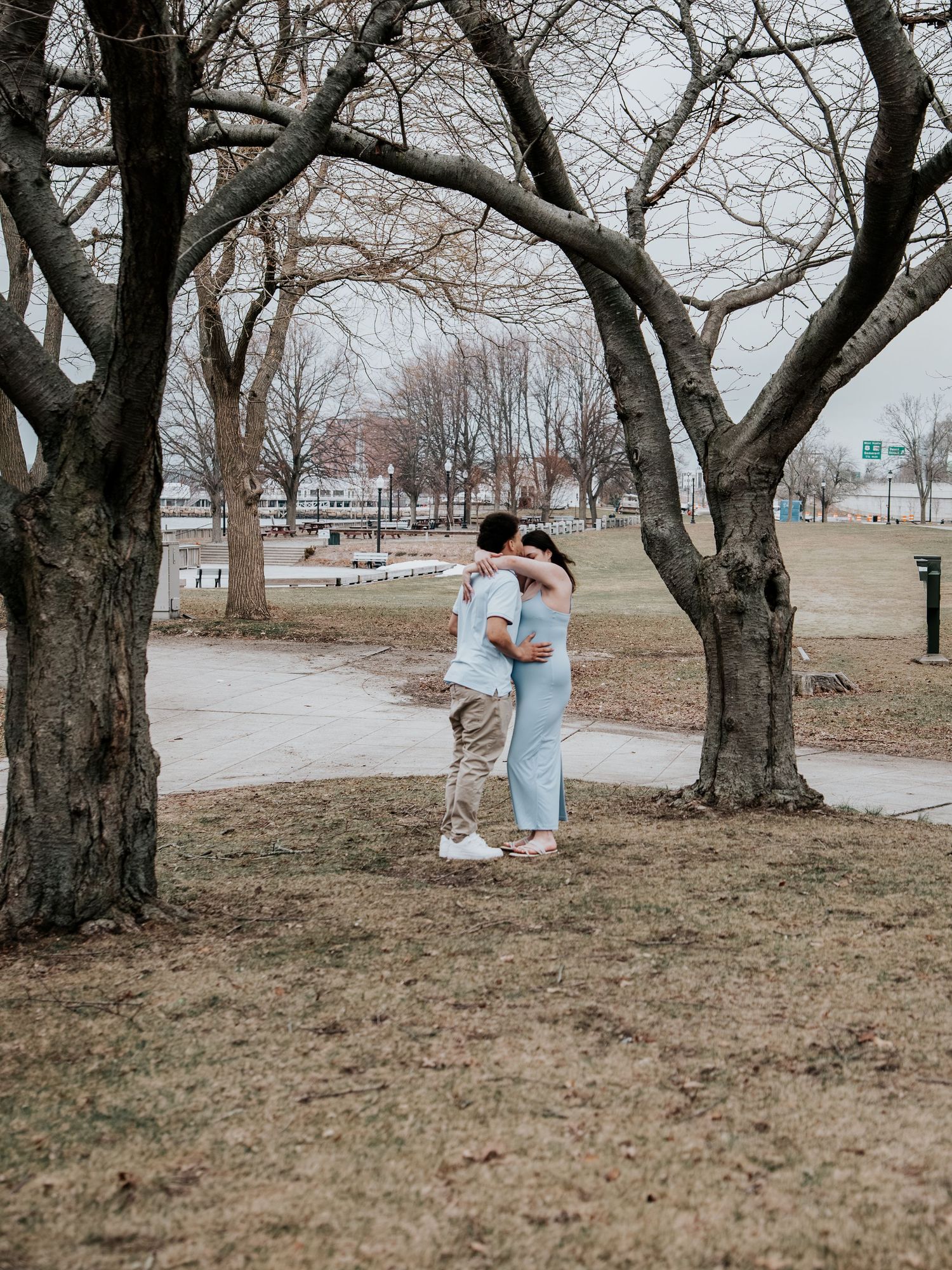 Couple embracing romantically in a park with bare trees and dry grass during early spring.