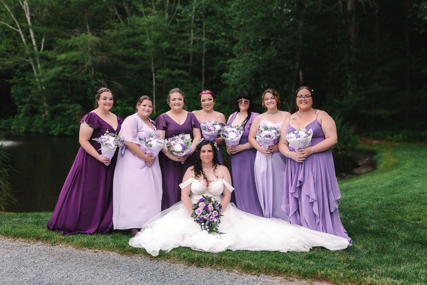 Bride in white gown seated with bridesmaids in purple dresses holding bouquets outdoors near a pond.
