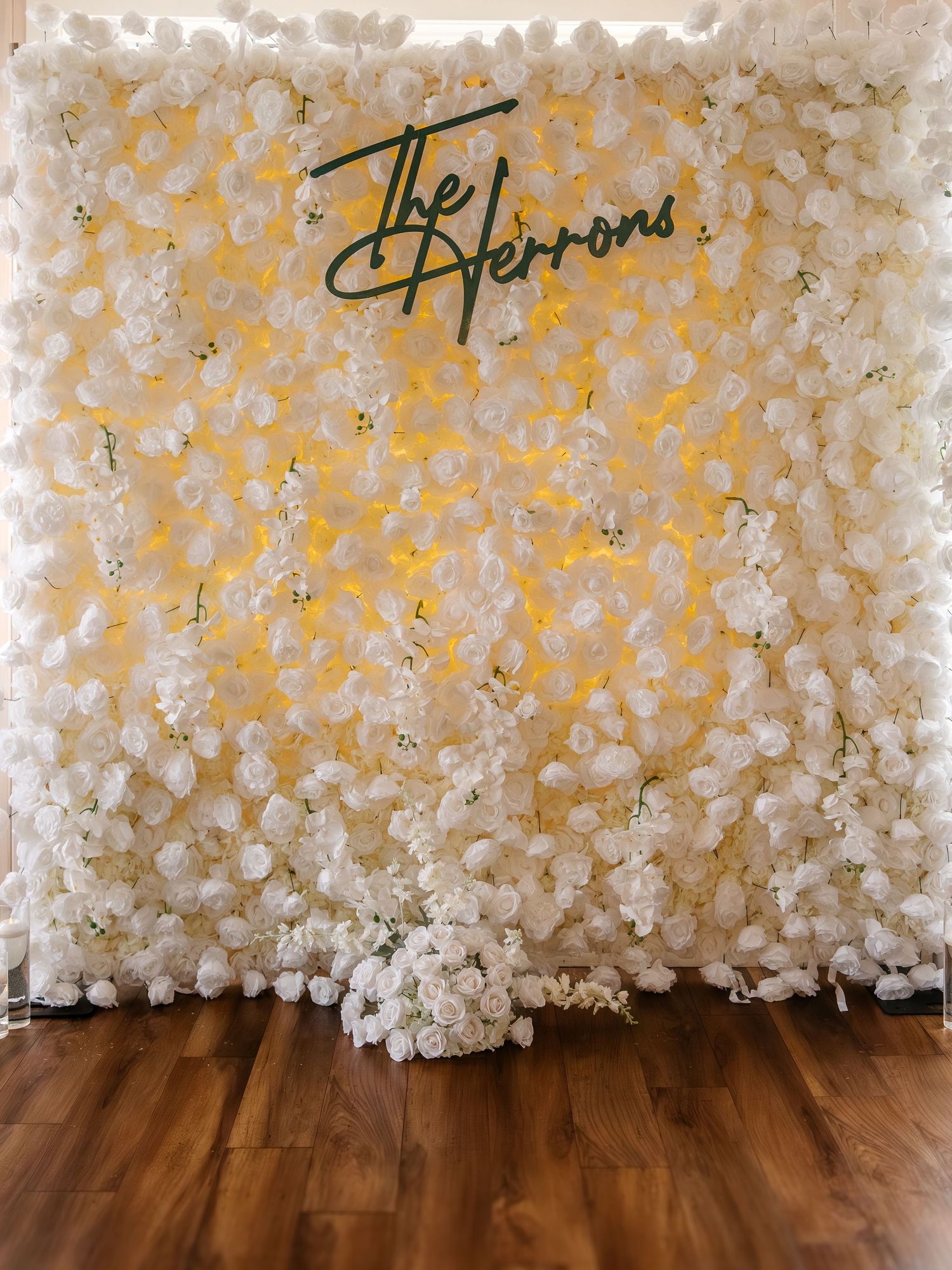 Elegant white flower wall backdrop with yellow accent lighting and green neon sign reading The Herrons on wood floor.