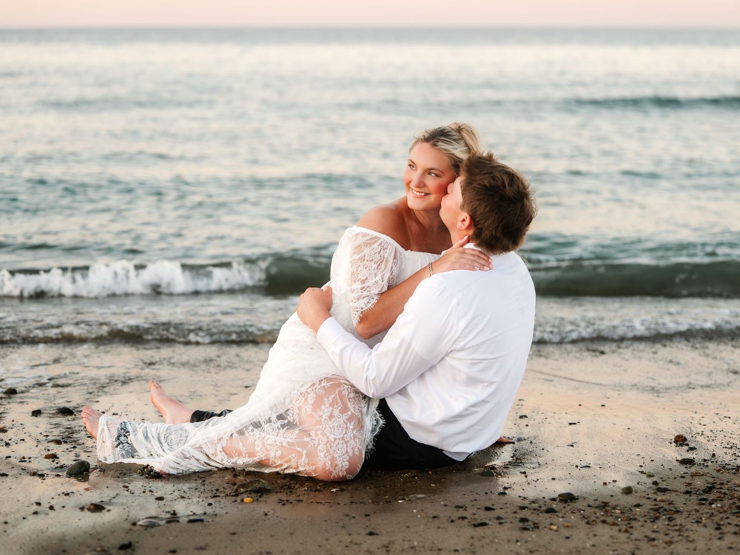 Couple in white wedding attire sitting on beach sand, embracing and smiling at sunset.