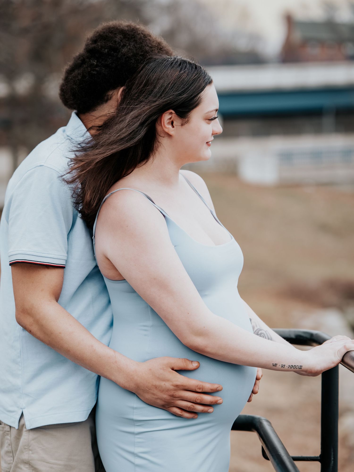 Couple sharing a tender maternity moment outdoors, woman in light blue dress cradling her pregnant belly by a railing.