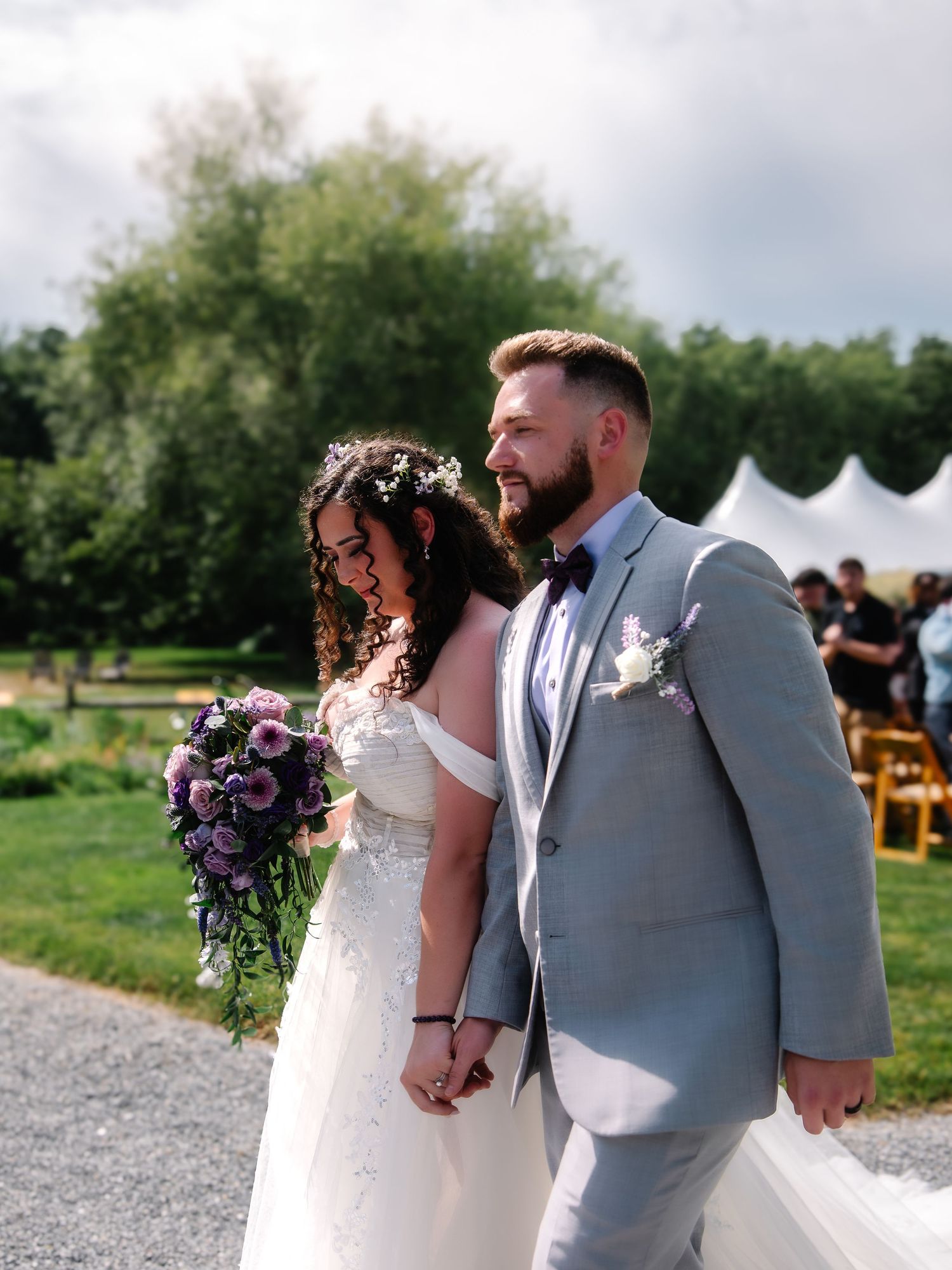 Bride and groom walk hand-in-hand after wedding ceremony, bride holding purple bouquet wearing floral crown.