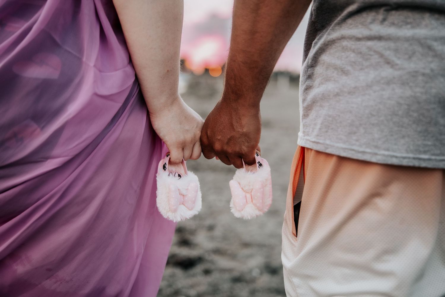 Couple holding hands and tiny pink baby booties on a beach, announcing pregnancy.