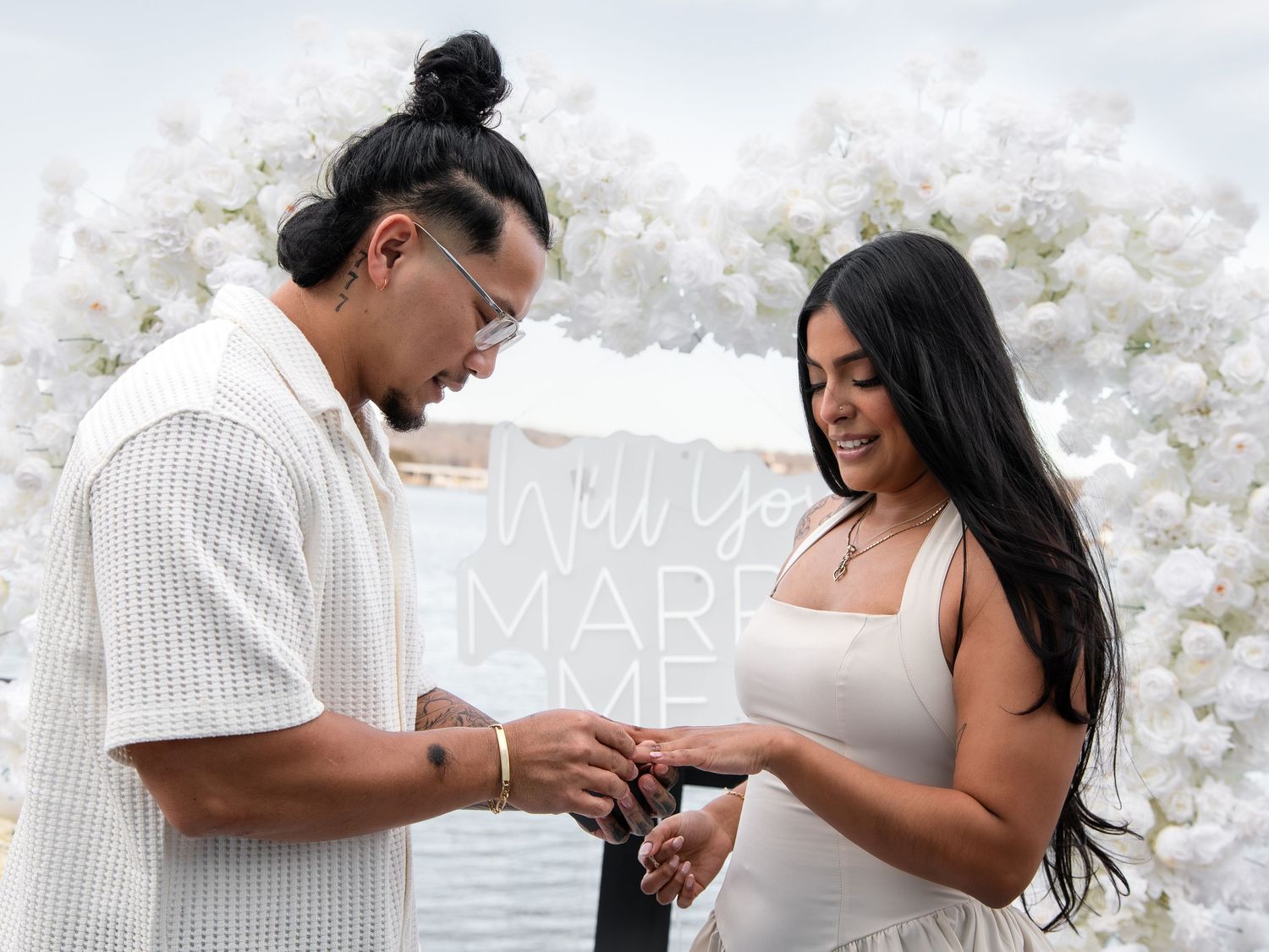 Couple sharing a romantic proposal moment outdoors with white floral arch and will you marry me sign.