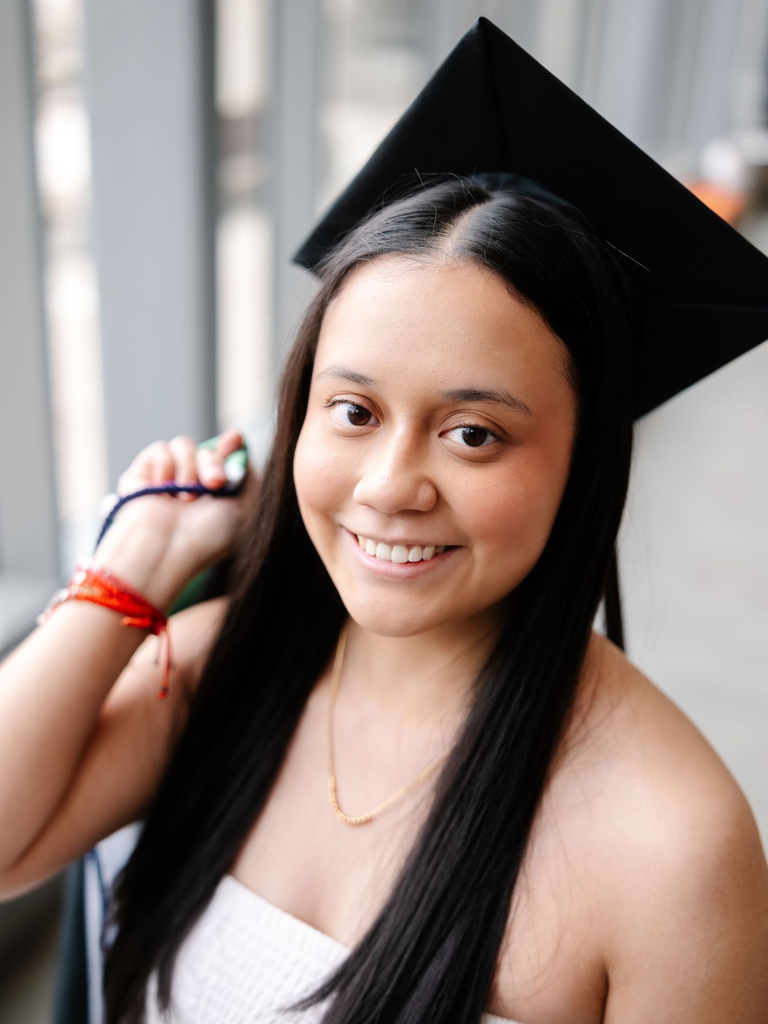 Smiling graduate in black mortarboard cap poses in white dress near bright window indoors.