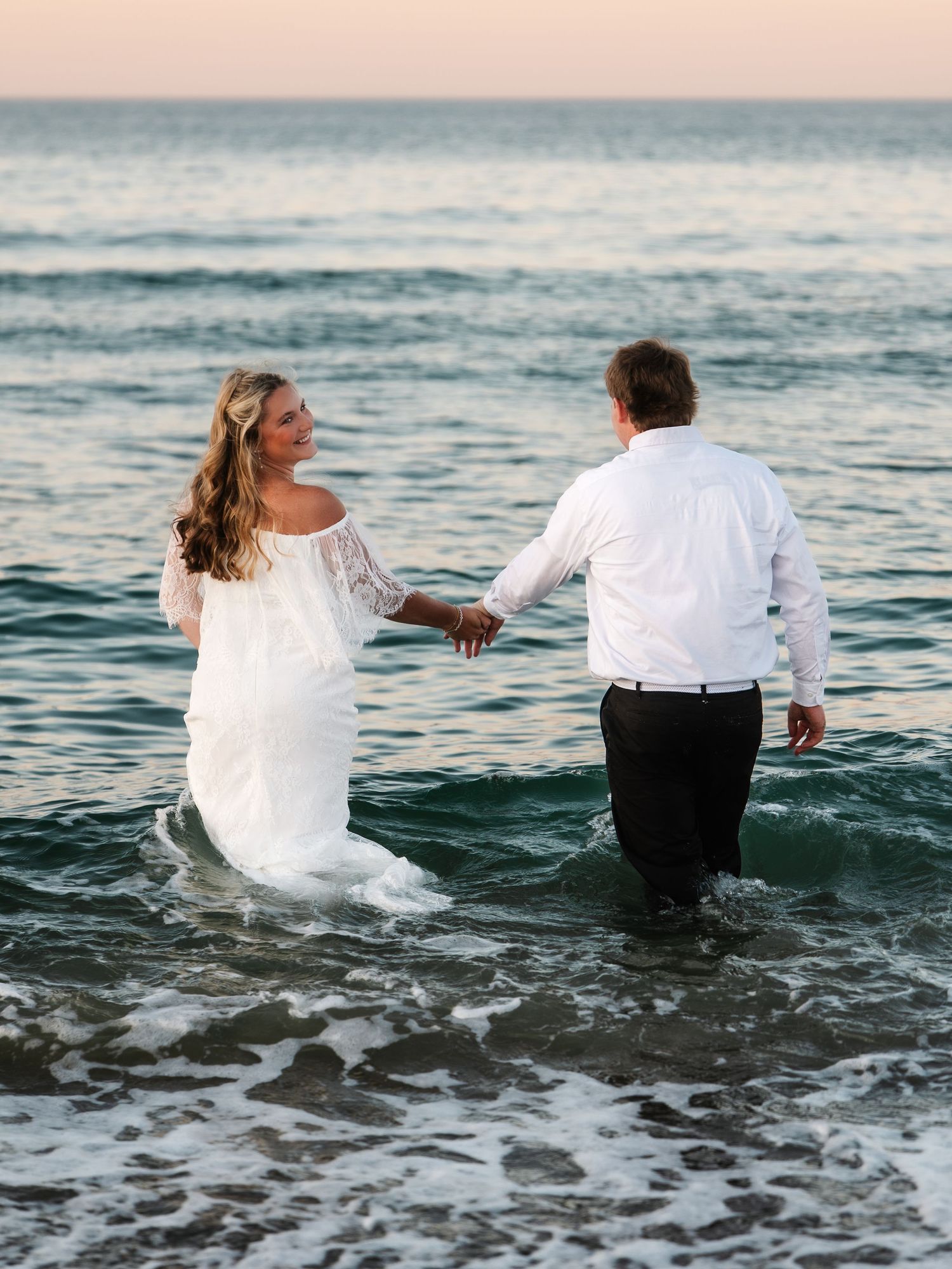 Bride and groom in wedding attire wading hand-in-hand through ocean waves at sunset beach.