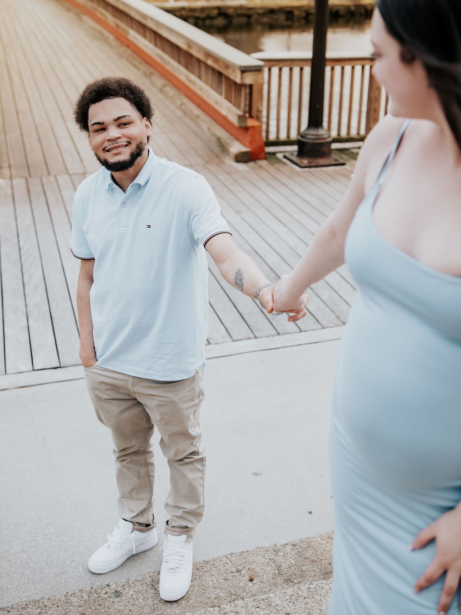 A smiling man in a light blue polo holds hands with a pregnant woman in a matching blue dress on a wooden boardwalk.
