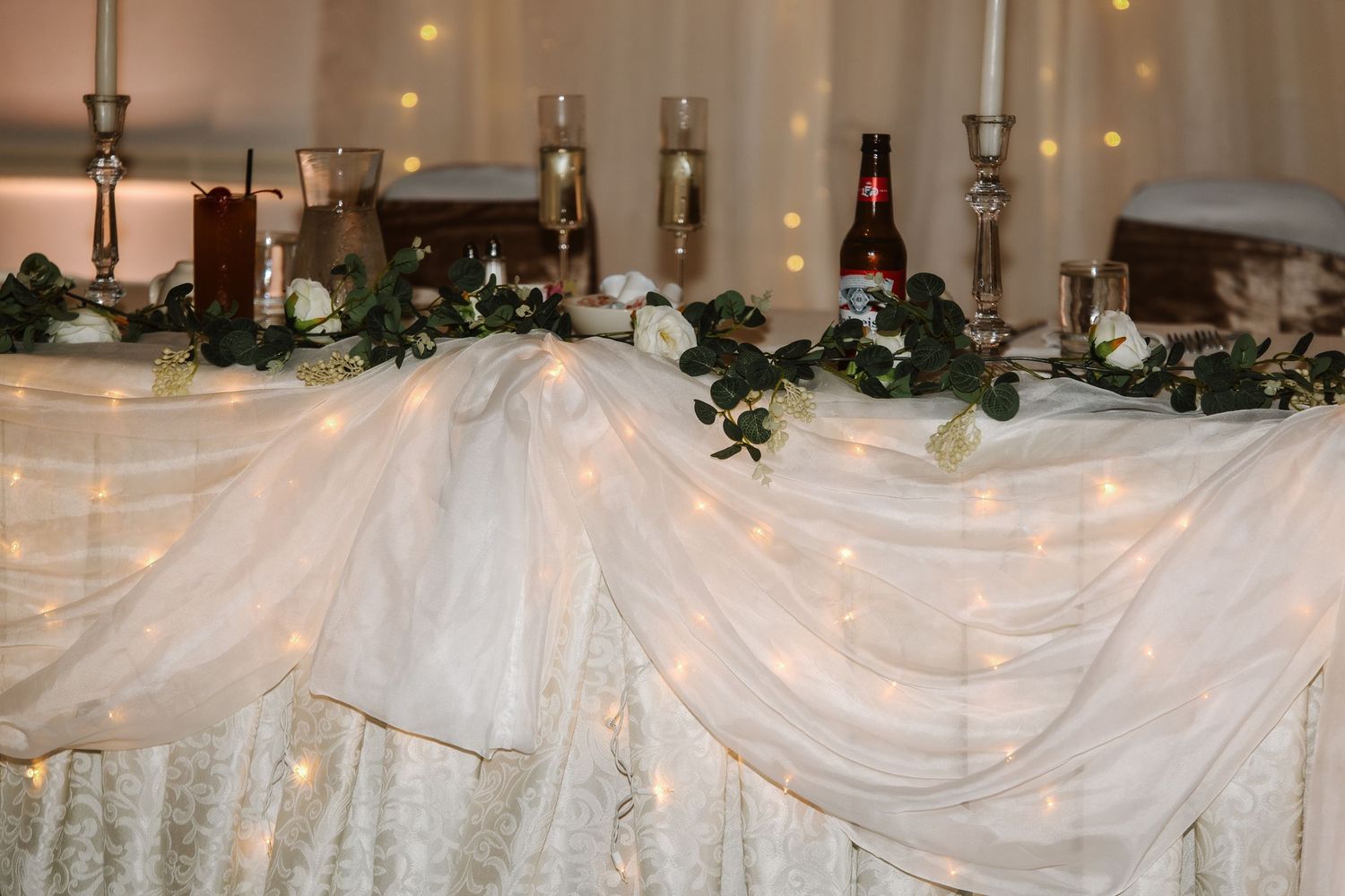 Elegant wedding head table with white tulle draping, fairy lights, floral greenery, candles, and champagne glasses.