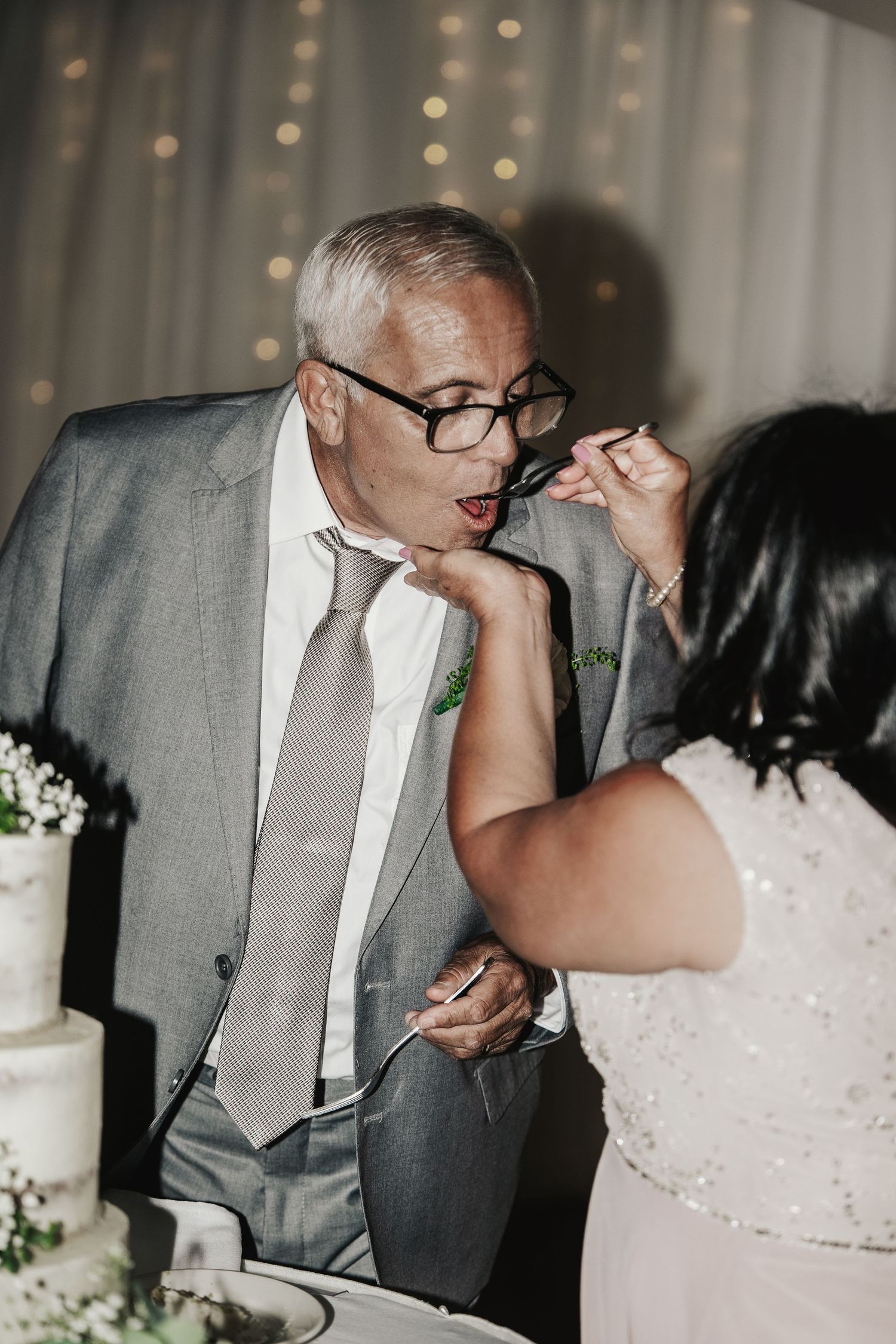 Groom in gray suit and glasses being fed wedding cake by bride at reception.