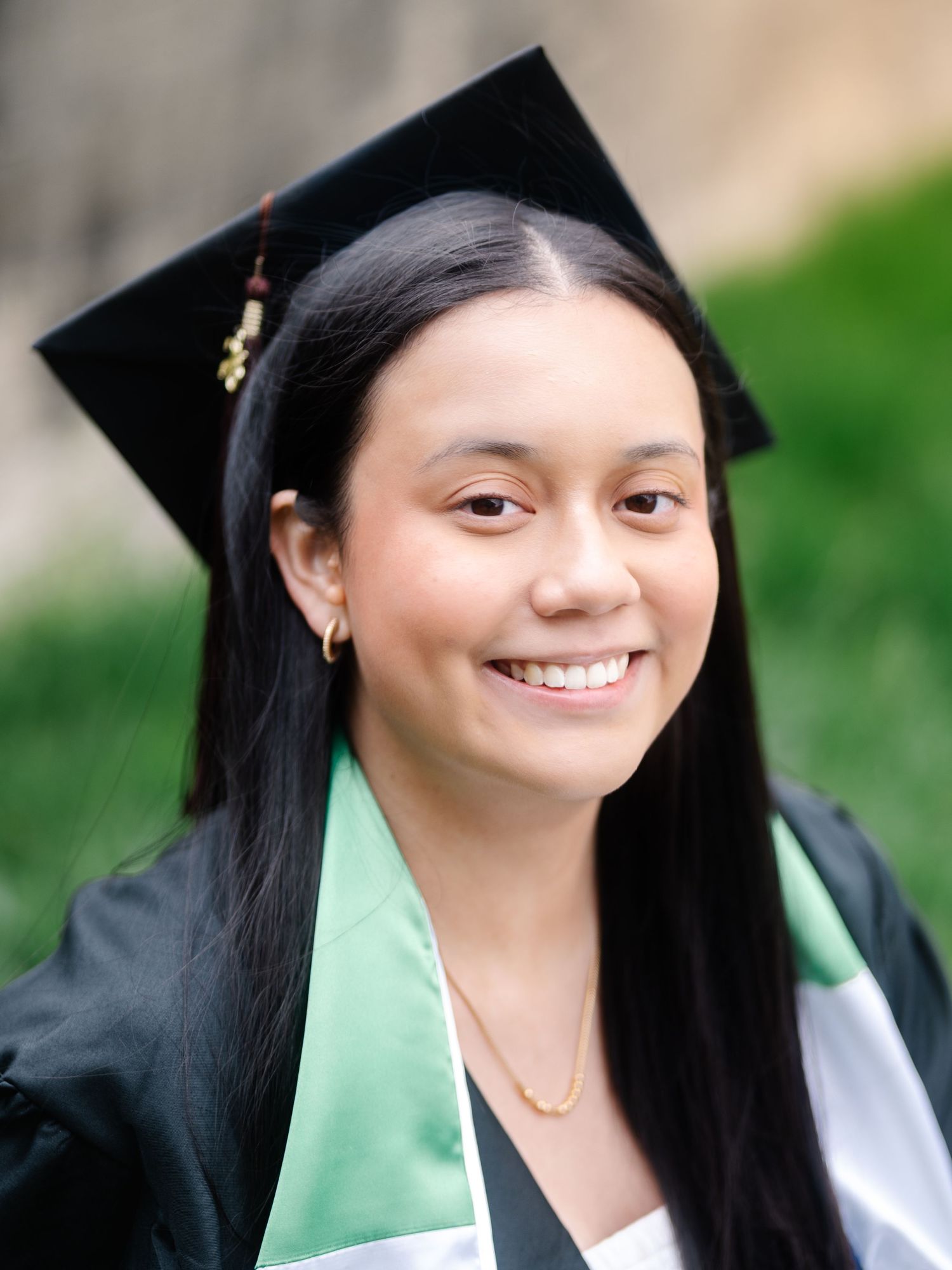 Smiling young woman in black graduation cap and gown with green honor stole outdoors.