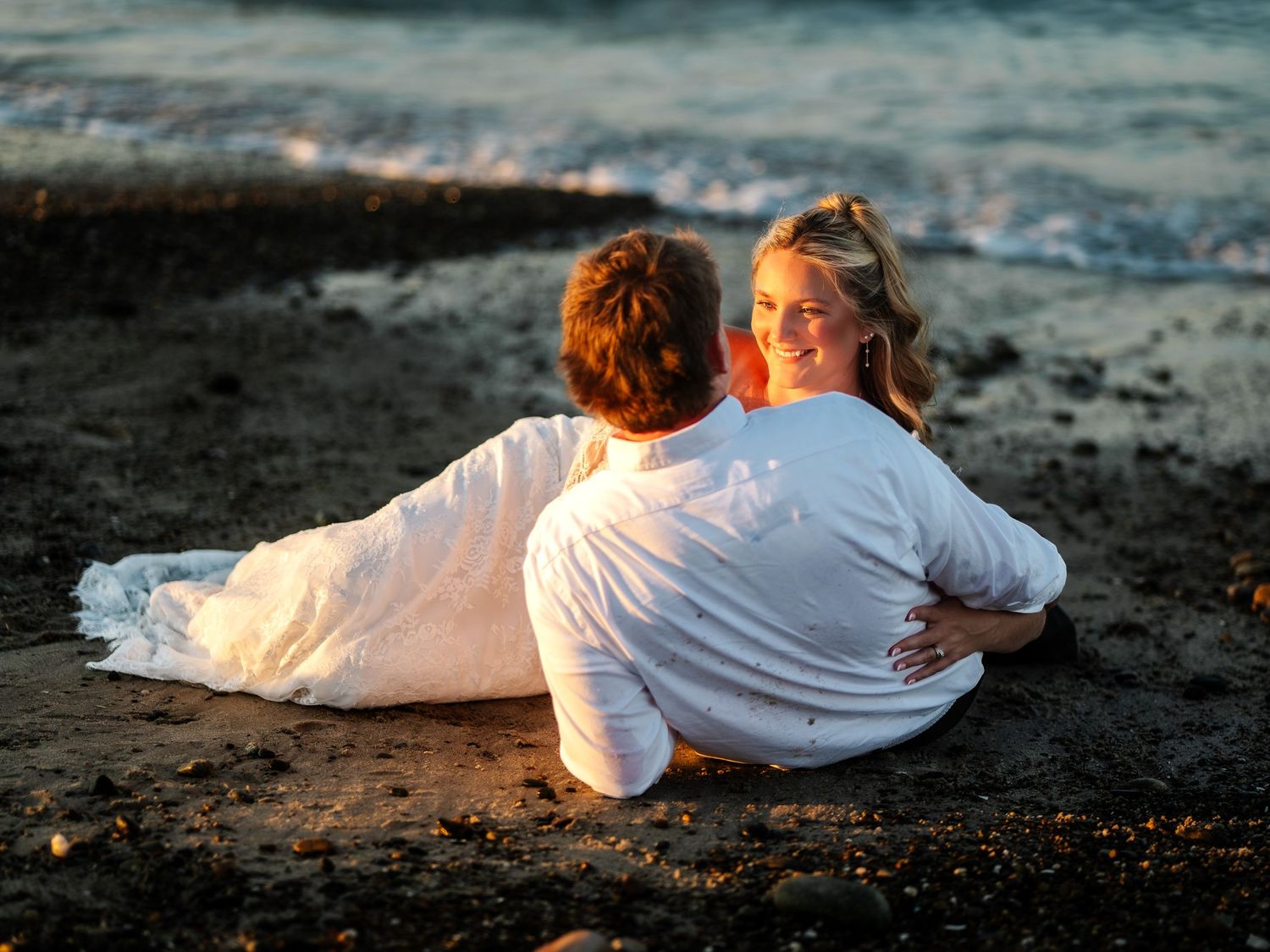 Happy couple relaxing together on sandy beach near ocean waves, woman in white dress smiling brightly.
