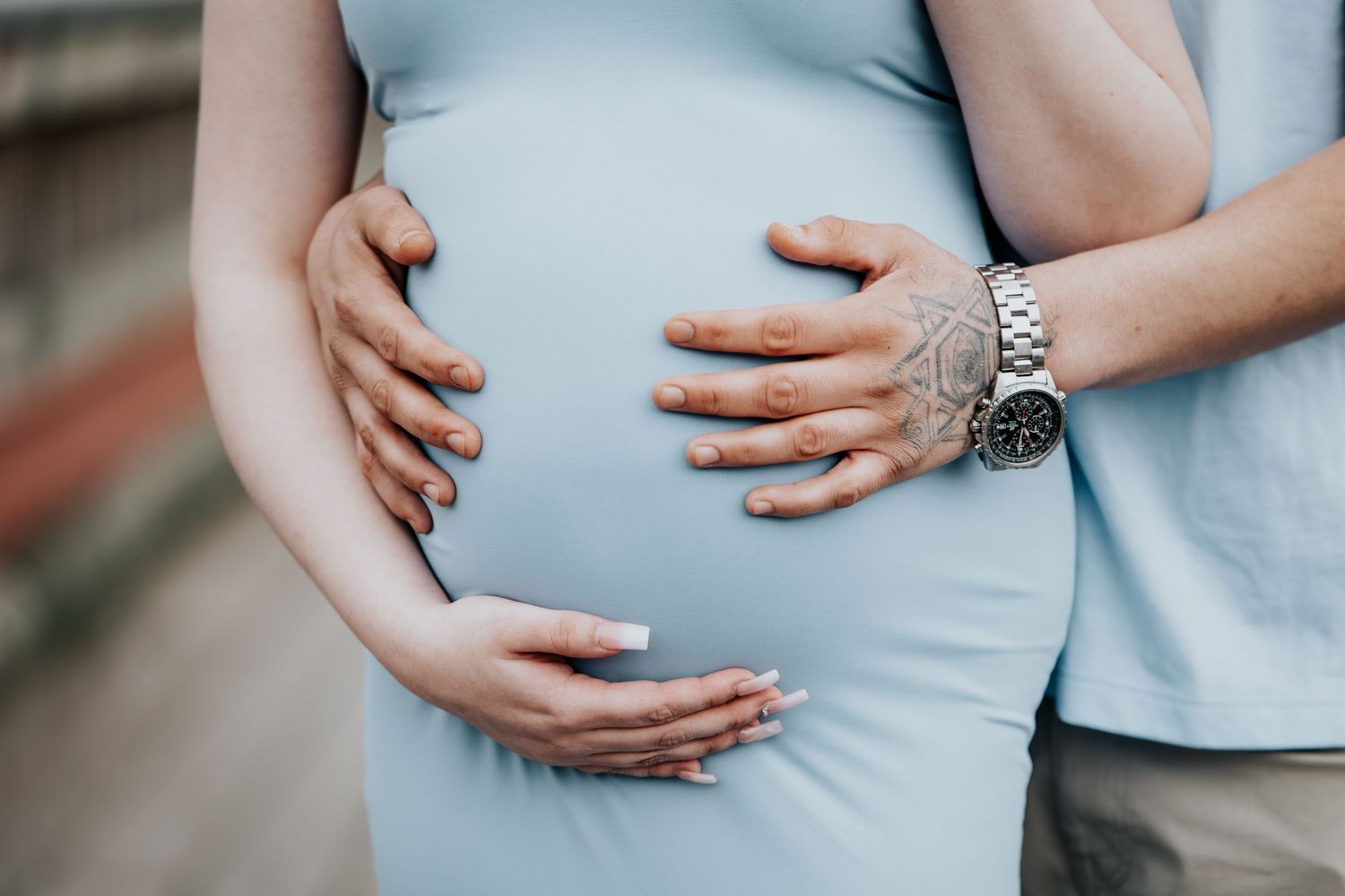Pregnant woman in blue dress with partner's hands gently cradling her baby bump, wearing a silver watch.