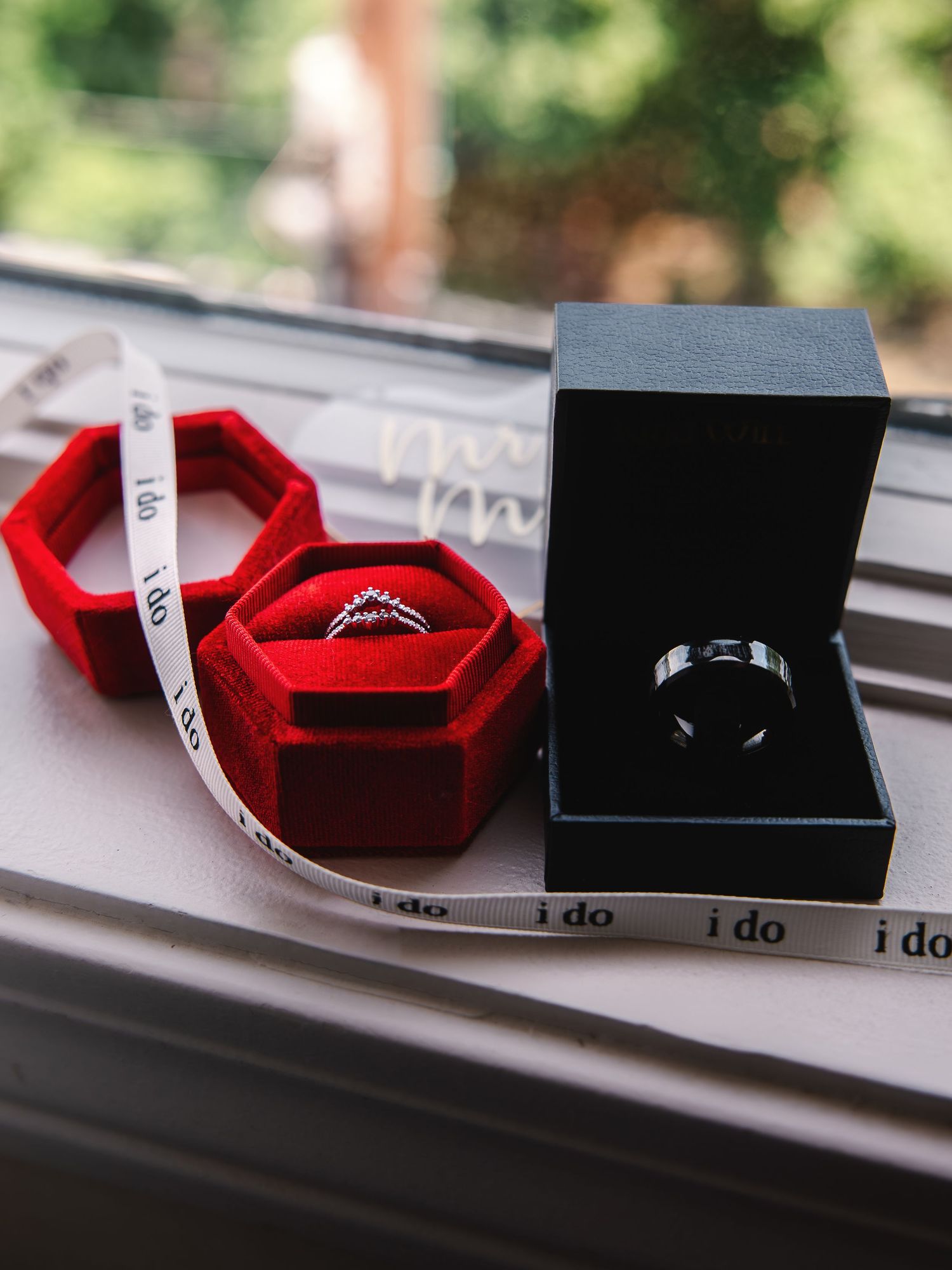 Two wedding rings displayed in elegant boxes with 'i do' ribbon, featuring a diamond ring in red velvet and a band in black box.