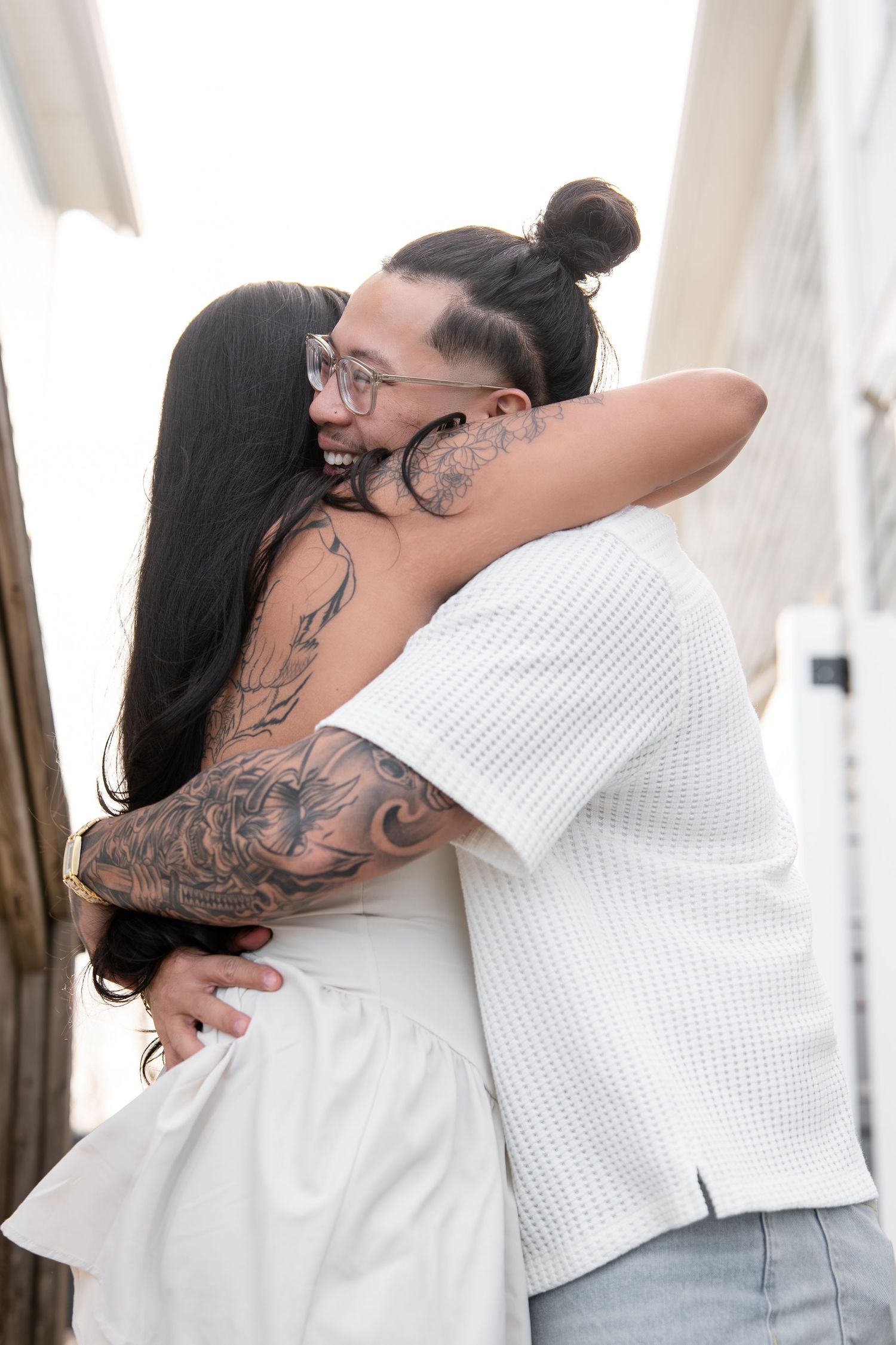 Couple sharing a warm embrace outdoors, woman with tattooed arm in white dress hugging man with bun hairstyle.