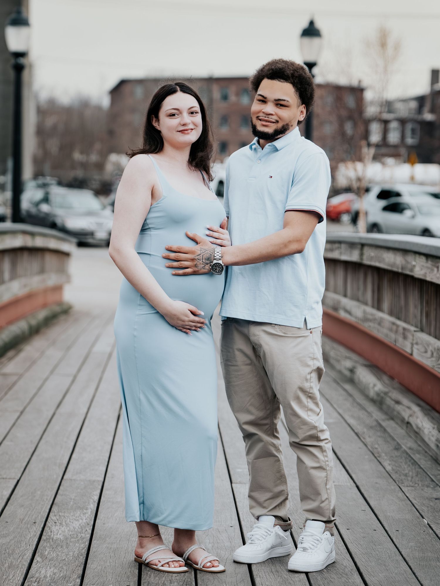 Pregnant couple posing on a wooden boardwalk, woman in light blue dress cradling baby bump, man in polo shirt beside her.
