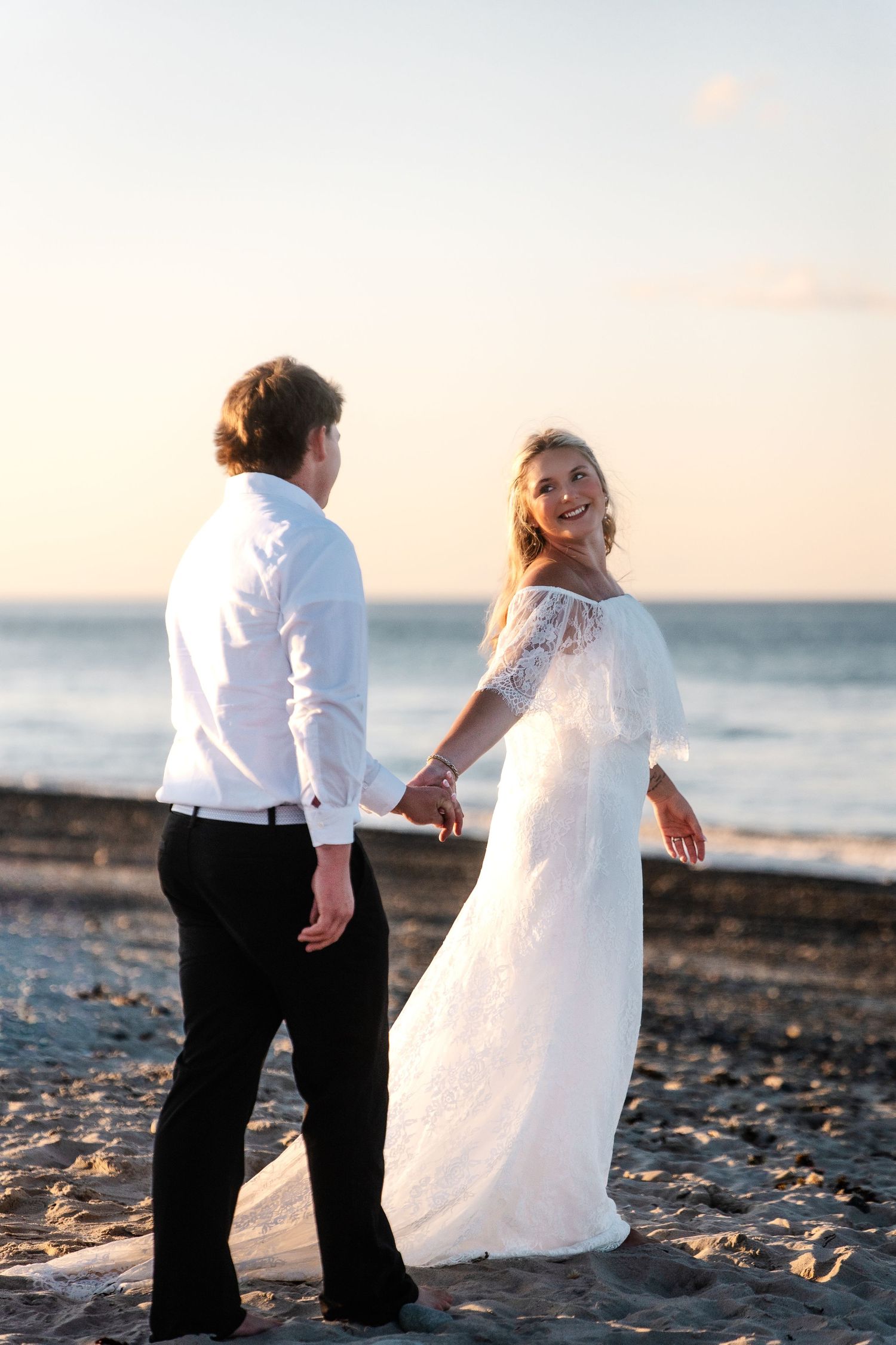 Smiling bride in white lace dress holding hands with groom while walking on beach at sunset.