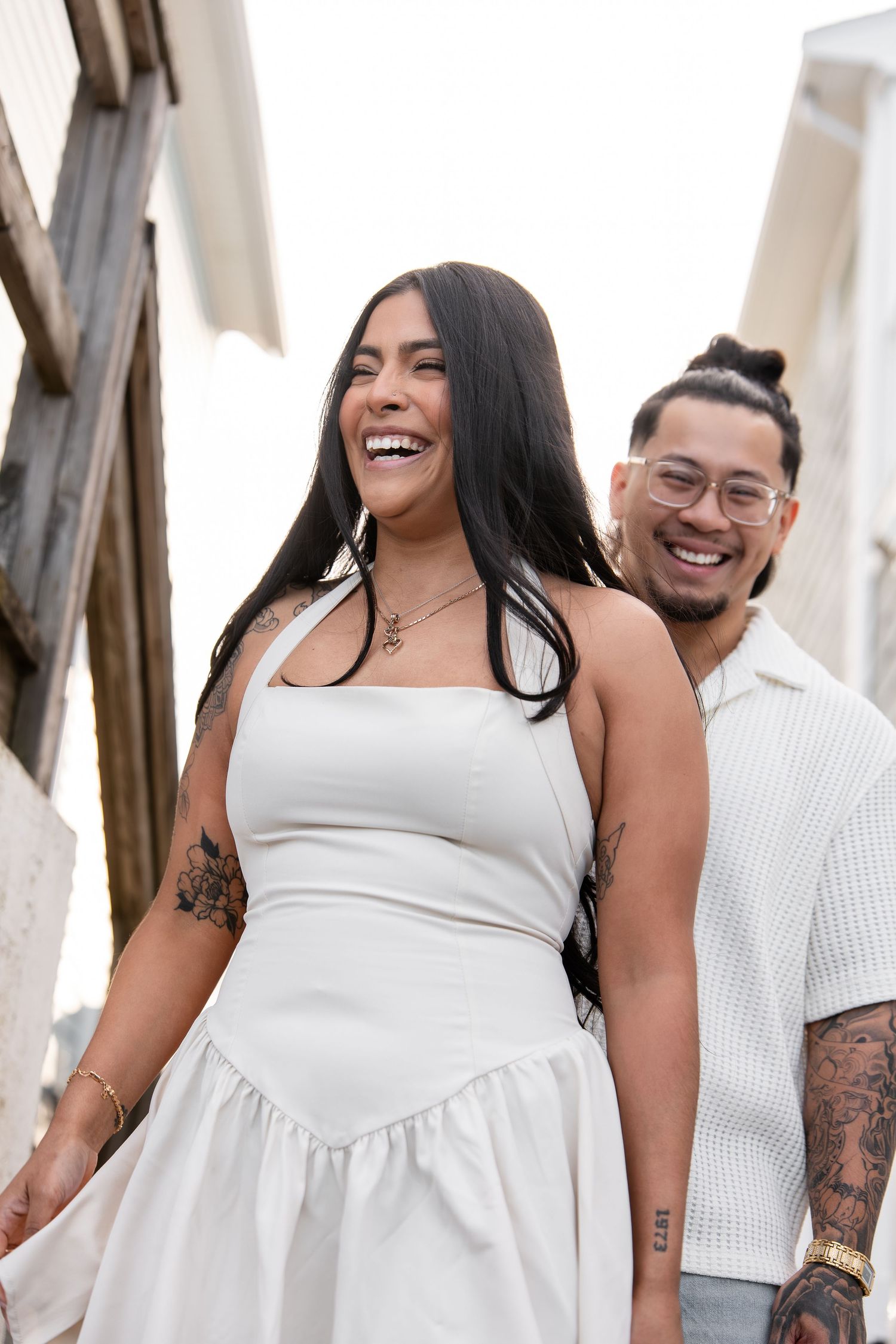 A laughing woman in a white dress with tattoos walks with a smiling man in glasses outdoors.