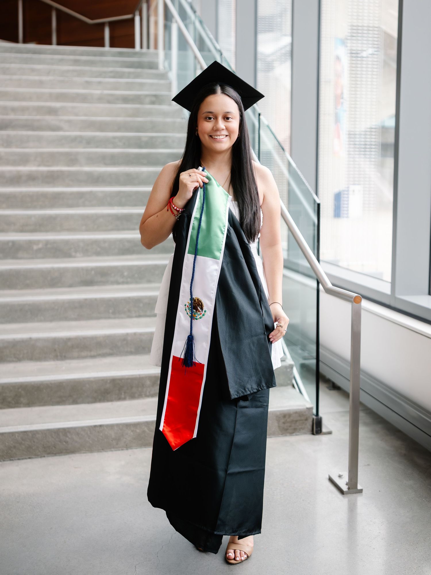 Graduate in black cap and gown holding Mexican flag stole, smiling on stairs at graduation ceremony.