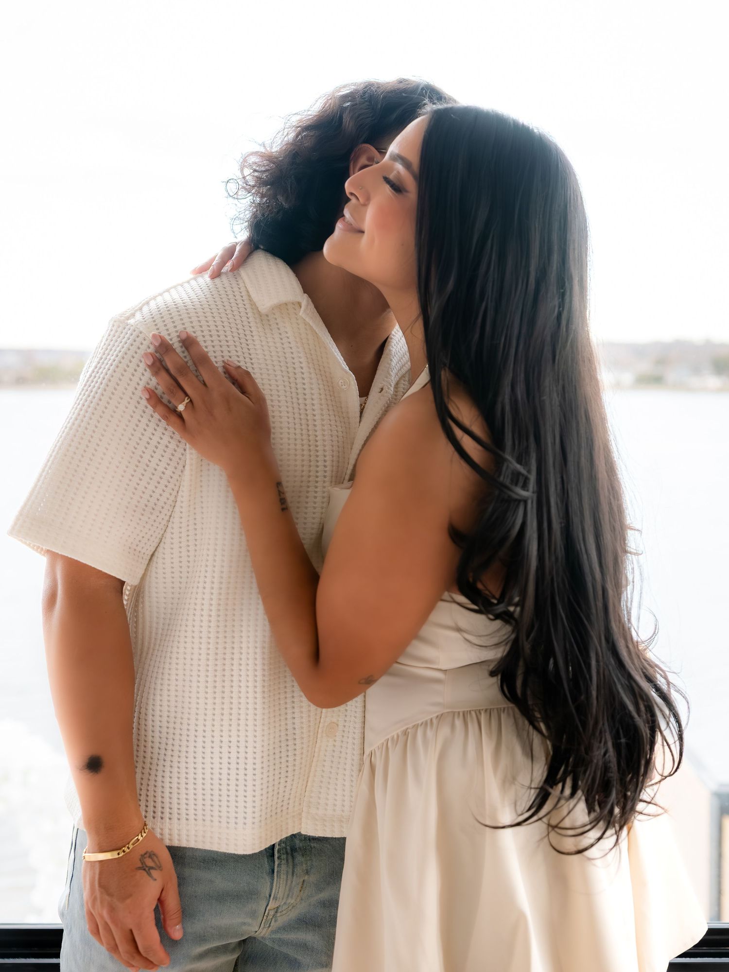 Couple hugging tenderly on a waterfront balcony, woman in white dress with long dark hair, man in cream shirt.