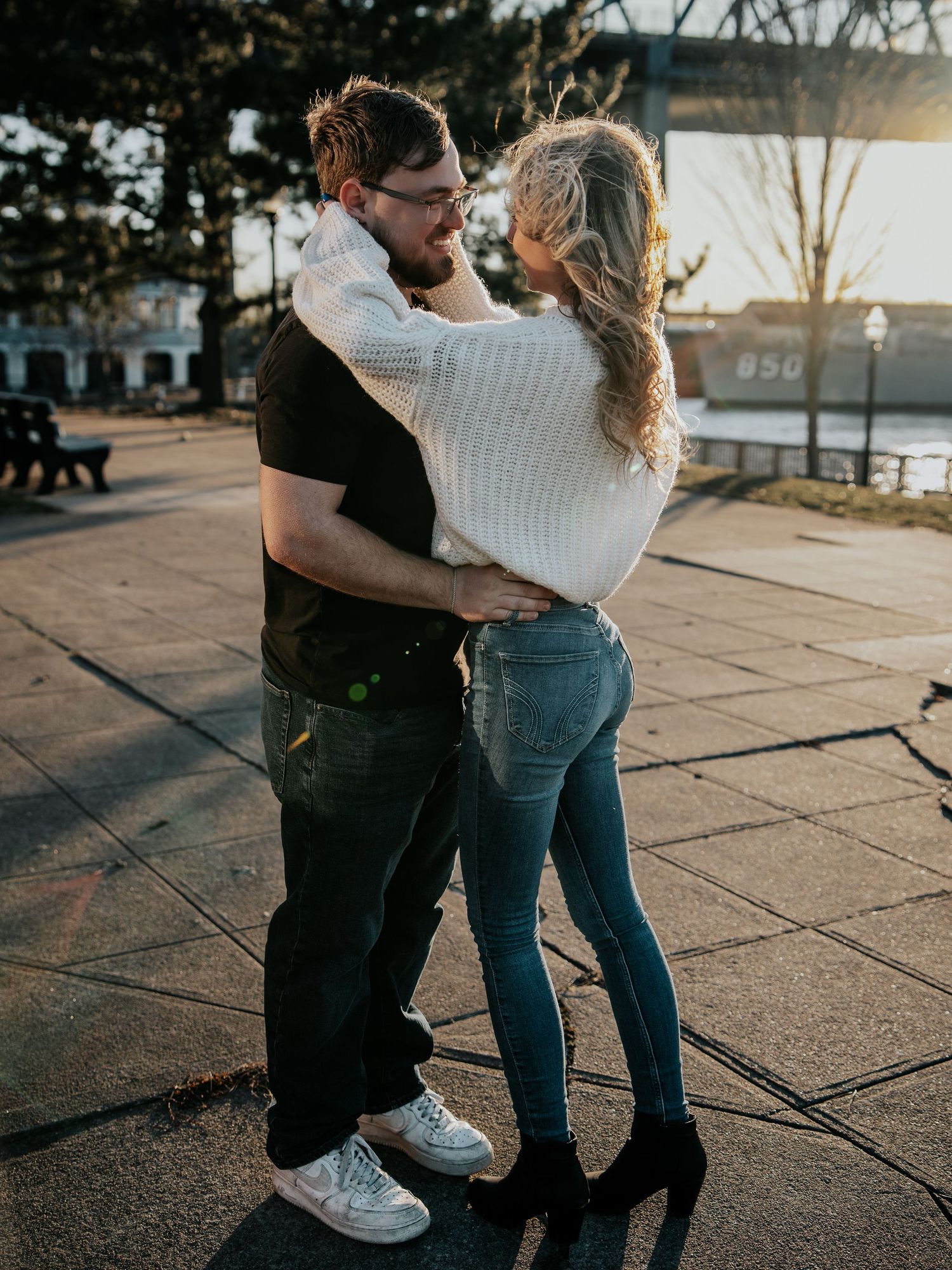 Romantic couple embracing outdoors at sunset near a waterfront, woman in white sweater and jeans, man in black tshirt.